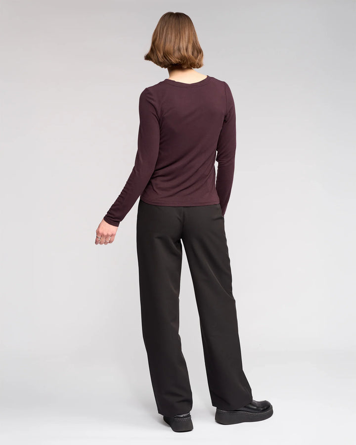 A person with short brown hair faces away, wearing the Knewe Label Hero Top in Raisin, black wide-leg pants, and black platform shoes against a plain light gray background.