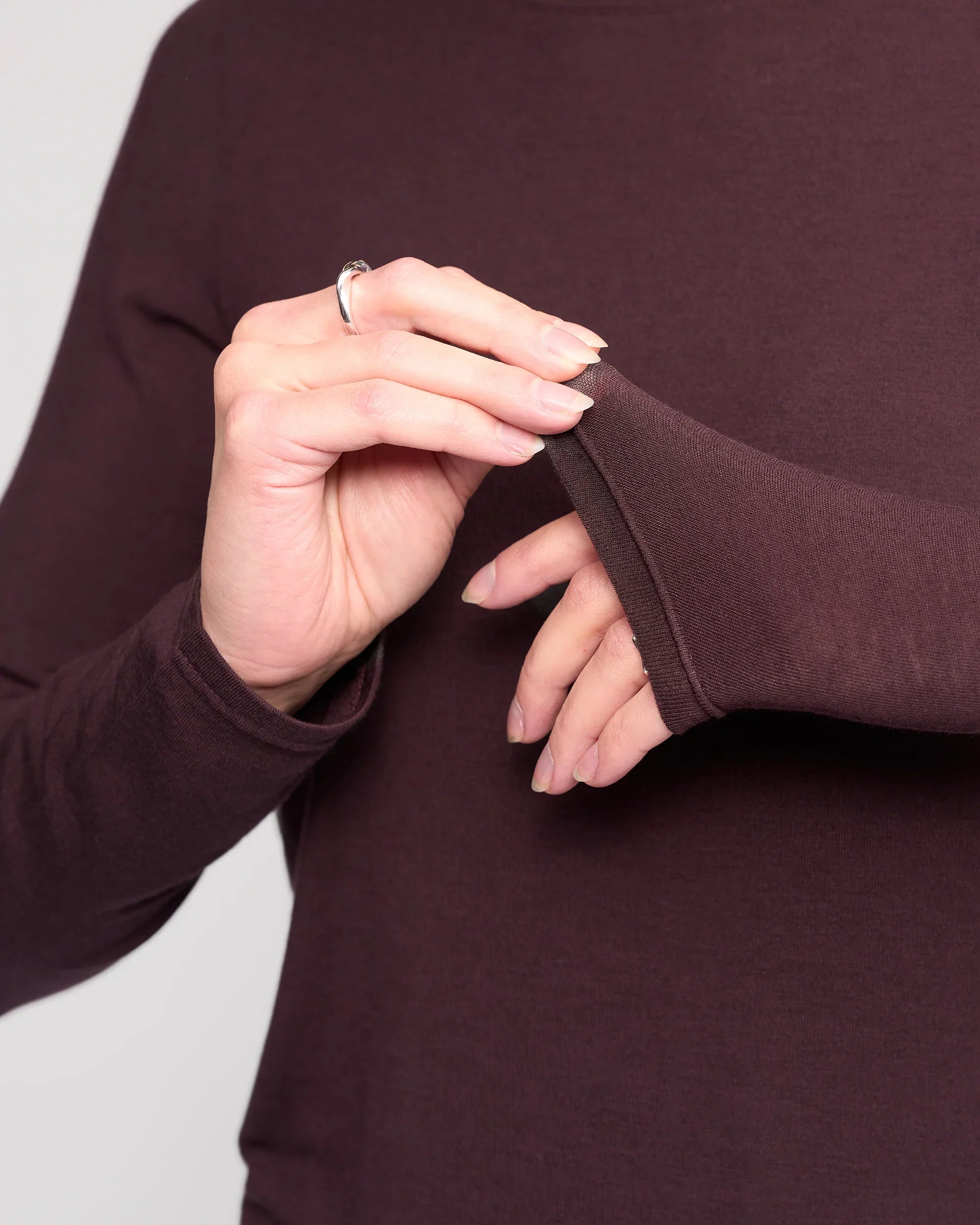 A person in the Knewe Label Hero Top – Raisin, a long-sleeve Tencel wool blend, gently pulls at the sleeve cuff to reveal their fingers and a silver ring against a neutral backdrop.