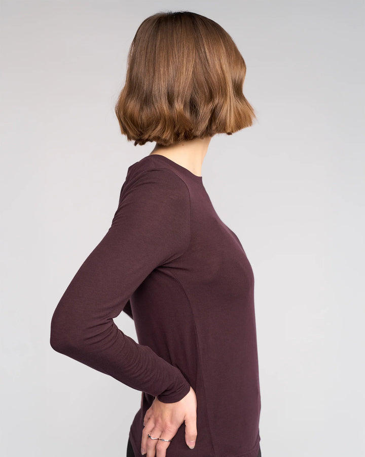 A person with chin-length light brown hair stands sideways, facing left, wearing Knewe Label's Hero Top in Raisin—a cozy Tencel wool blend—resting one hand on their hip against a plain light background.