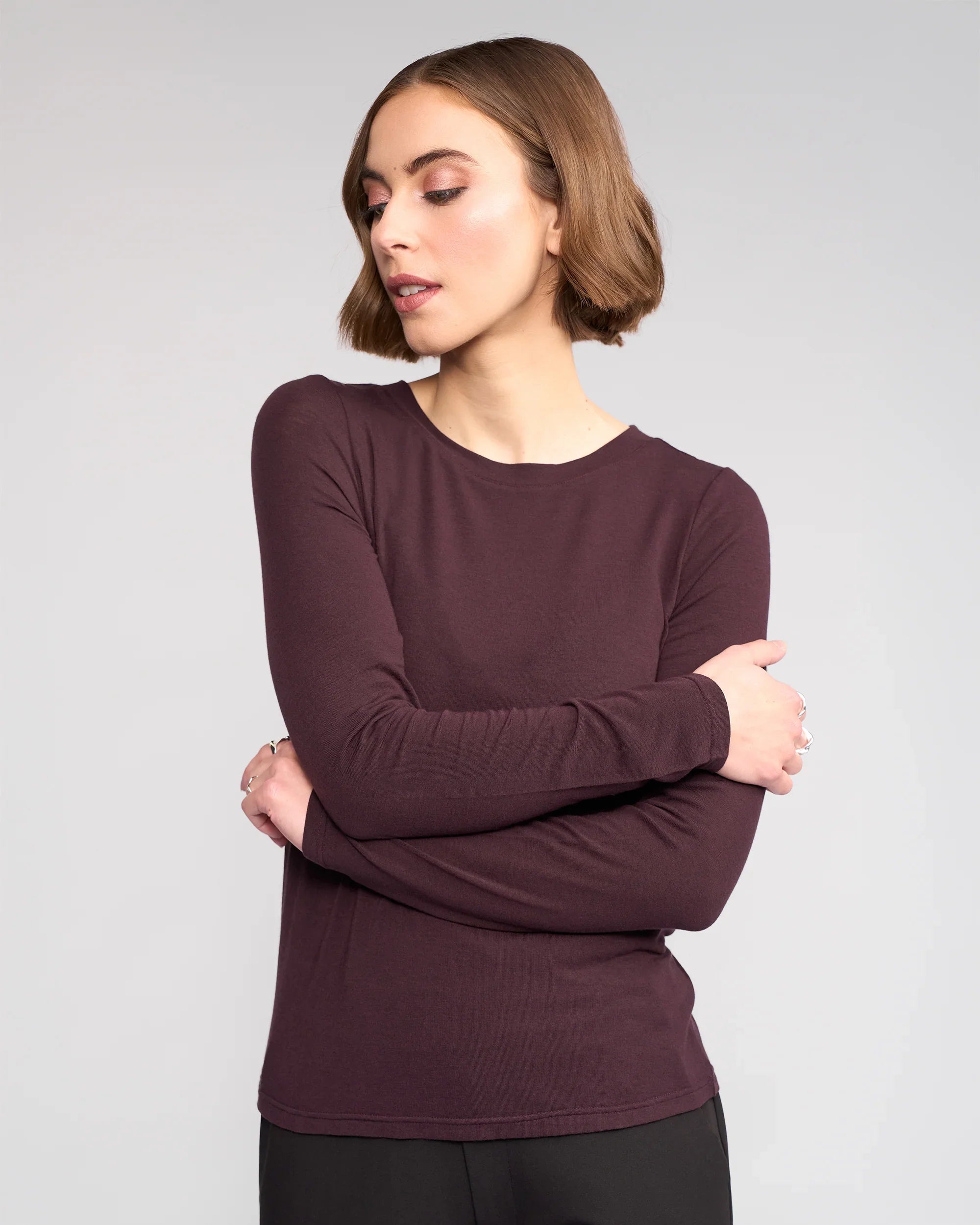 A woman with short brown hair crosses her arms and looks to the side, wearing the long-sleeved Knewe Label Hero Top in Raisin against a plain light background.