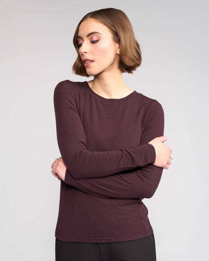 A woman with short brown hair crosses her arms and looks to the side, wearing the long-sleeved Knewe Label Hero Top in Raisin against a plain light background.