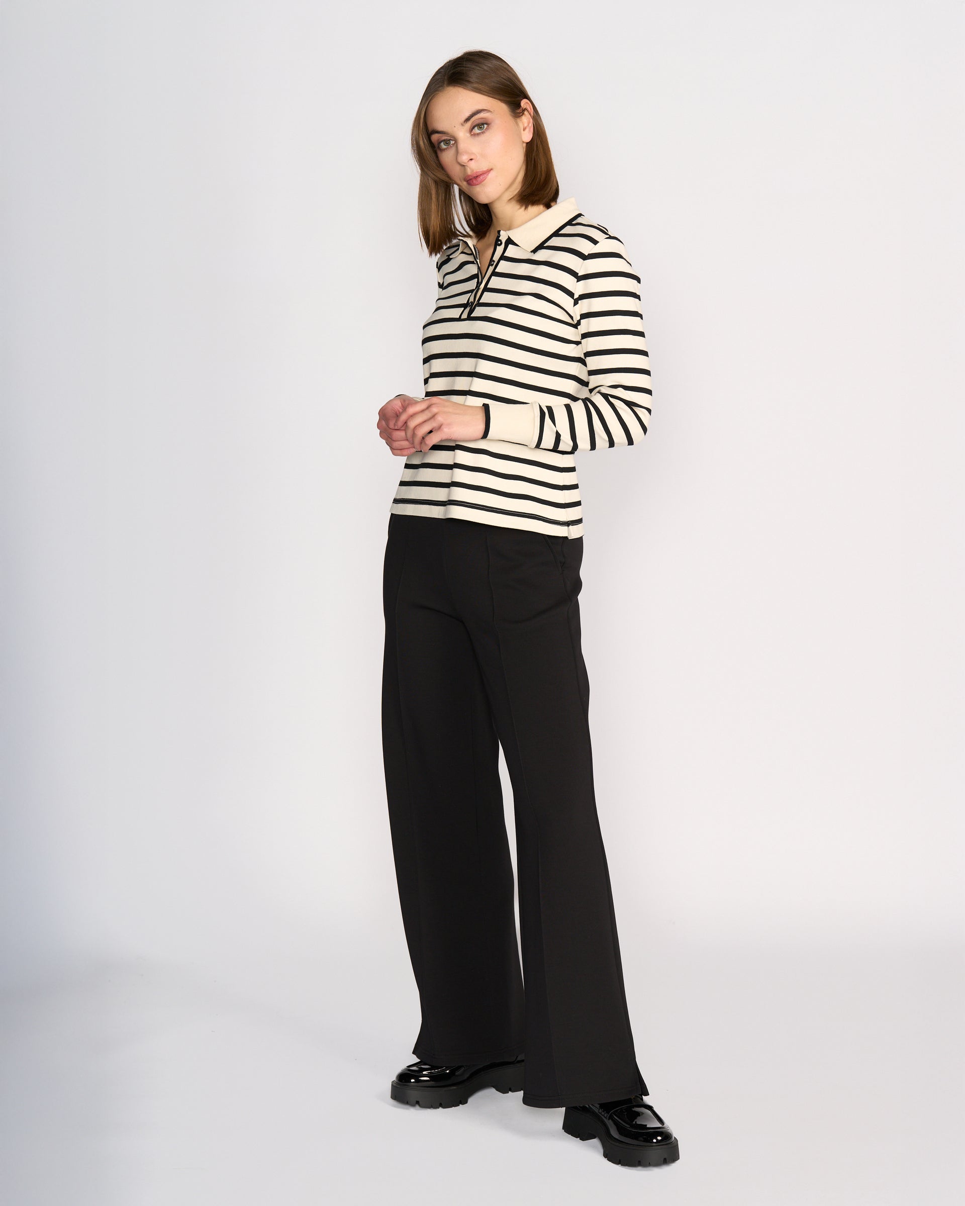A woman stands against a white background wearing the Knewe Label Era Top in oat and black stripes, paired with black wide-leg pants and shiny shoes. The soft cotton top has long sleeves, and she looks to the side with straight, shoulder-length hair.