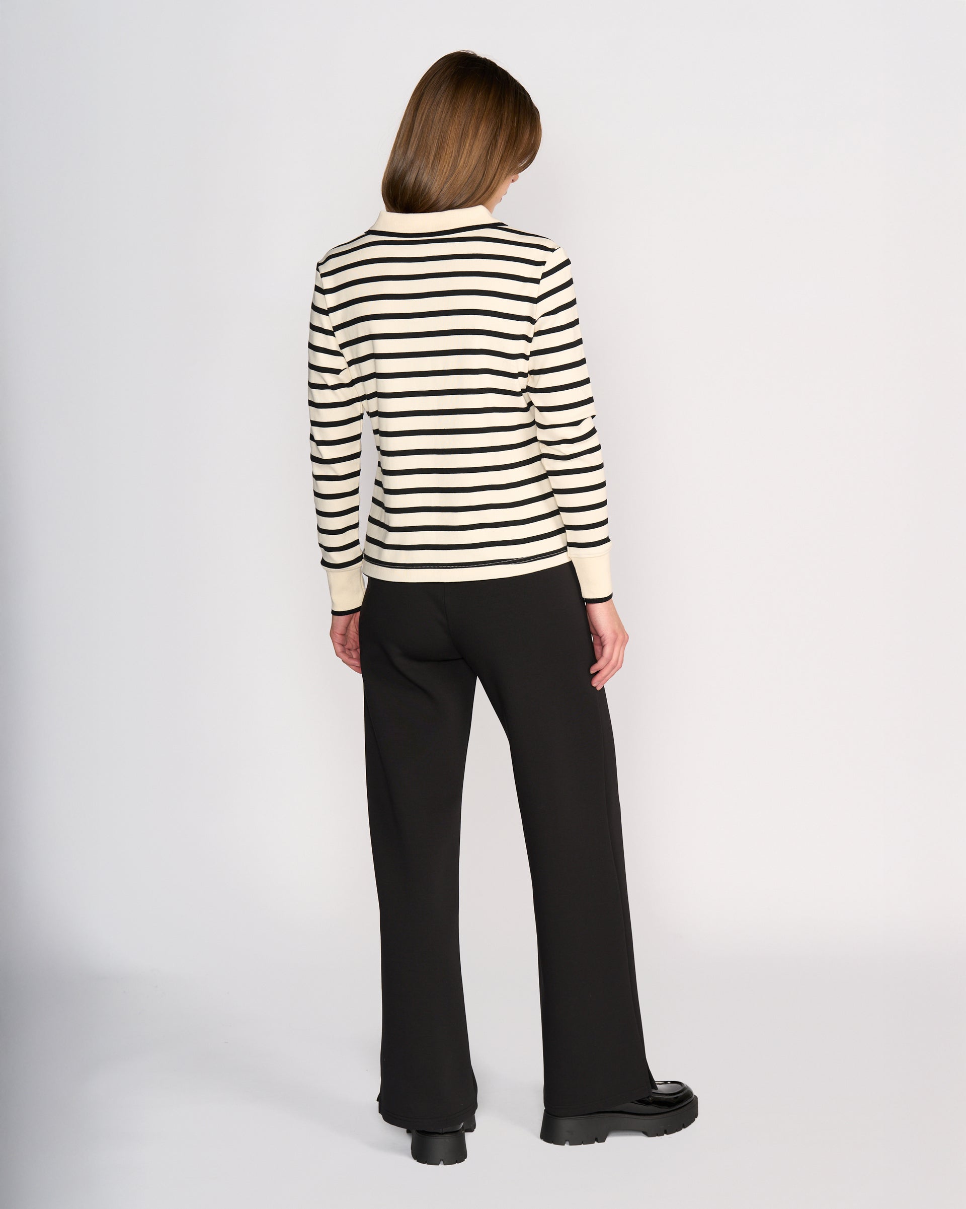 A person with straight, shoulder-length hair stands facing away, wearing the Knewe Label Era Top in oat/black stripes paired with loose black pants and black shoes, set against a plain white background.
