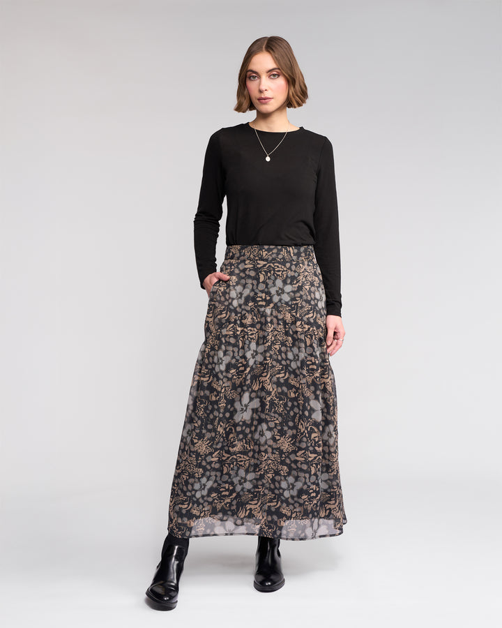 A woman with short light brown hair stands against a plain background, wearing the Knewe Label Milan Skirt - Hazy, a gold-green floral full-length skirt with an elasticated waist, paired with a black top, black boots, and a pendant necklace. Her hands rest in her pockets.