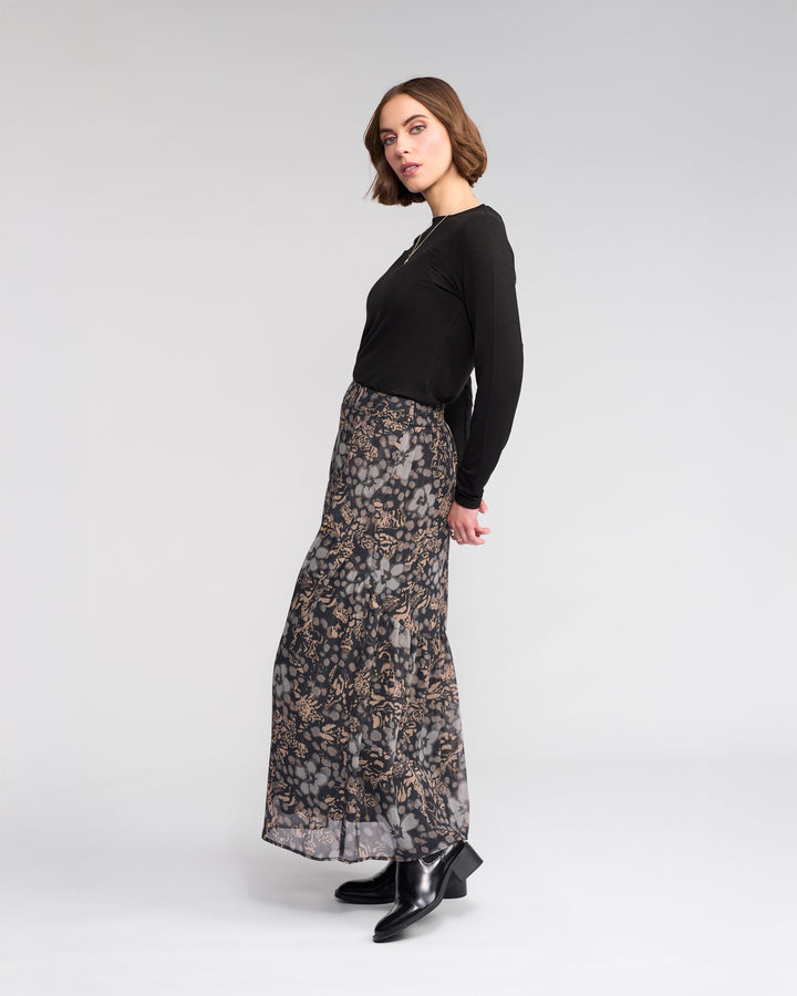 A woman with short brown hair stands sideways in a studio, wearing the Knewe Label Milan Skirt - Hazy, a black long-sleeve top, and black ankle boots against a light gray background.
