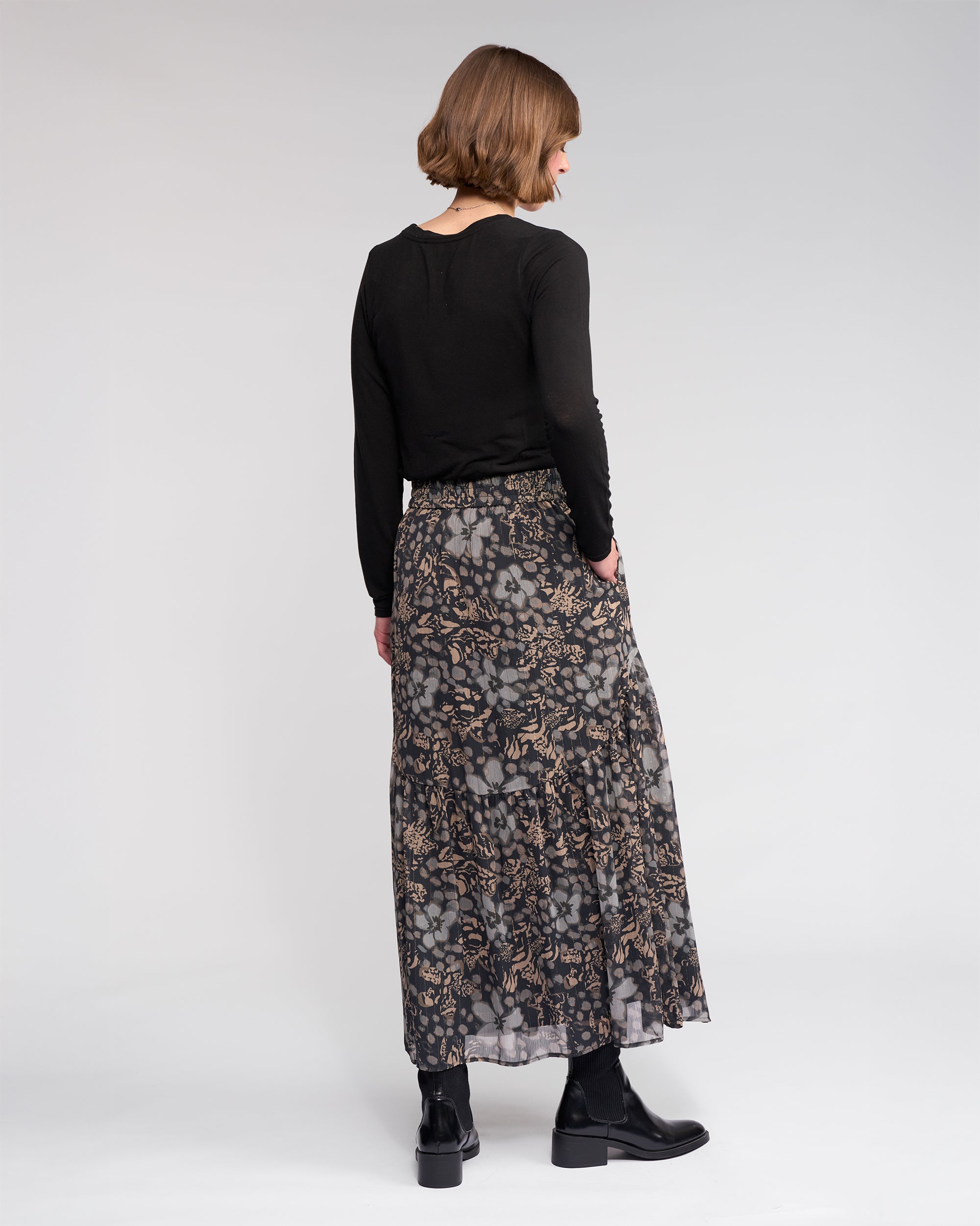 A woman with short light brown hair faces away from the camera, wearing a black long-sleeve top, black ankle boots, and the Milan Skirt - Hazy by Knewe Label against a plain light background.