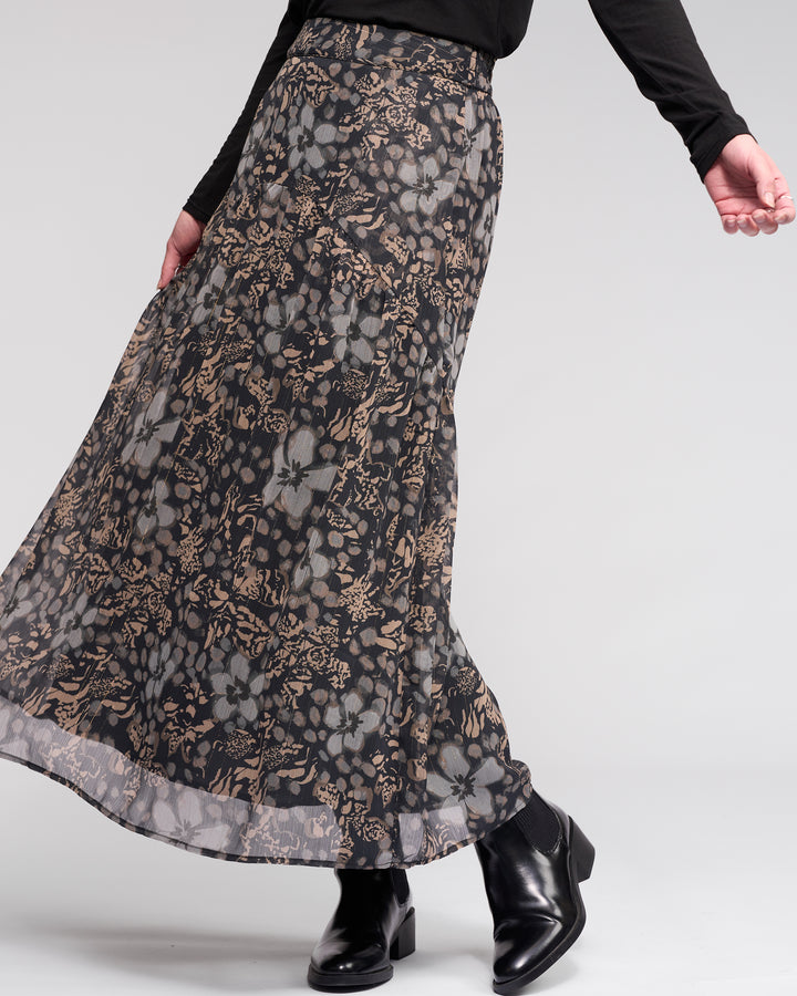 Someone stands against a light gray background wearing the Knewe Label Milan Skirt - Hazy, a long, flowy gold-green floral skirt with an elasticated waist, paired with a black long-sleeve top and black ankle boots.