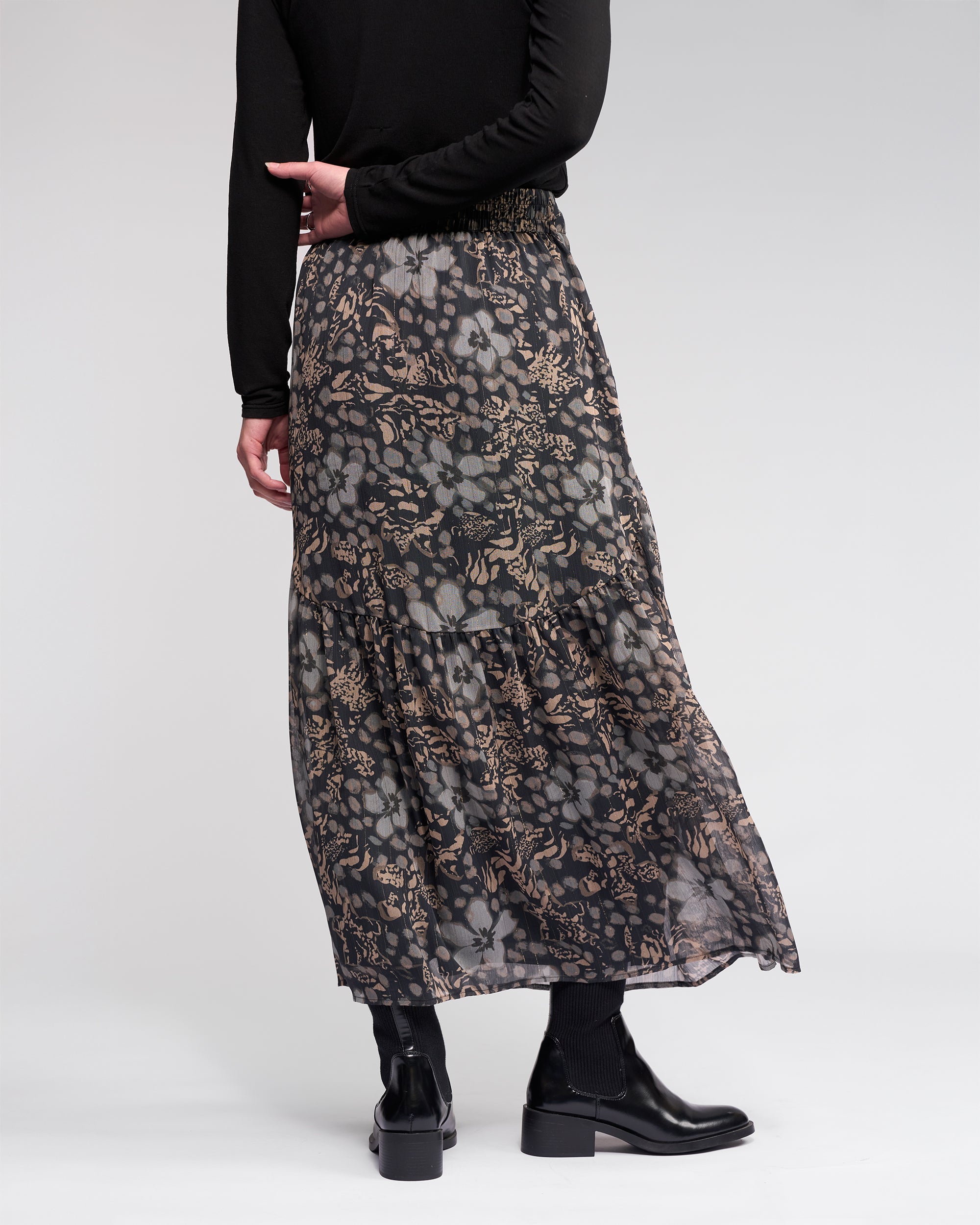 A person wearing the Knewe Label Milan Skirt - Hazy, a full-length sheer floral skirt in black and beige with an elasticated waist, paired with a black top and ankle boots, stands facing away against a plain light background.
