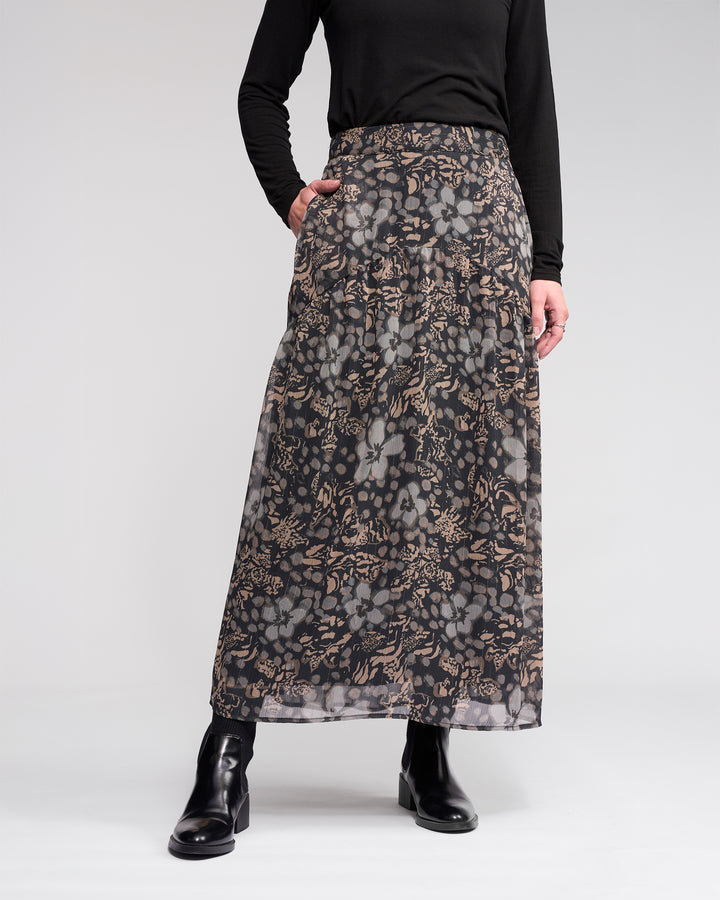 A person wearing the Knewe Label Milan Skirt - Hazy, a black long-sleeve top, and black ankle boots stands with one hand in their pocket against a plain light background.
