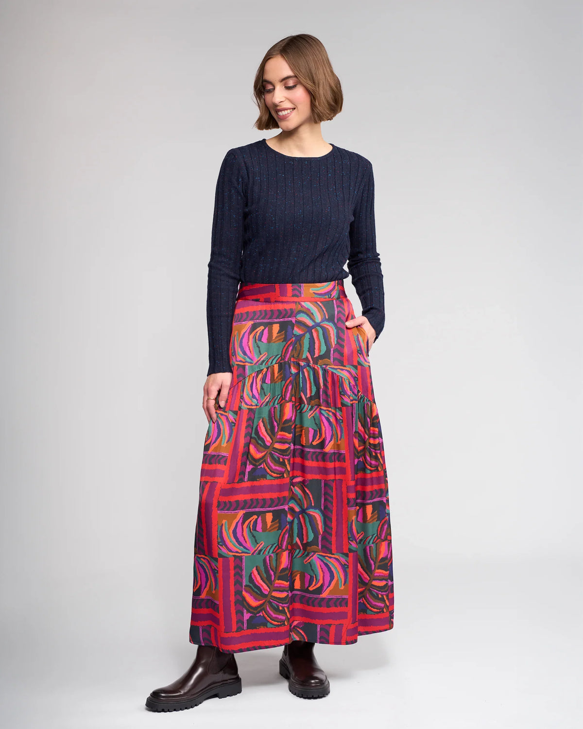 A woman smiles with one hand in her pocket, wearing the Knewe Label Milan Skirt - Sunset, featuring red and purple tones, paired with a navy long-sleeve sweater and dark ankle boots against a plain background.
