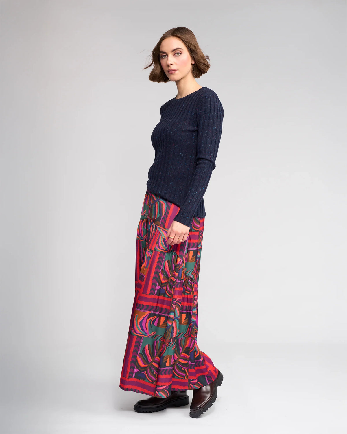 A woman with short brown hair stands sideways against a plain light gray background, wearing a fitted navy ribbed sweater, Knewe Label’s Milan Skirt - Sunset in red and green, and brown chunky shoes.