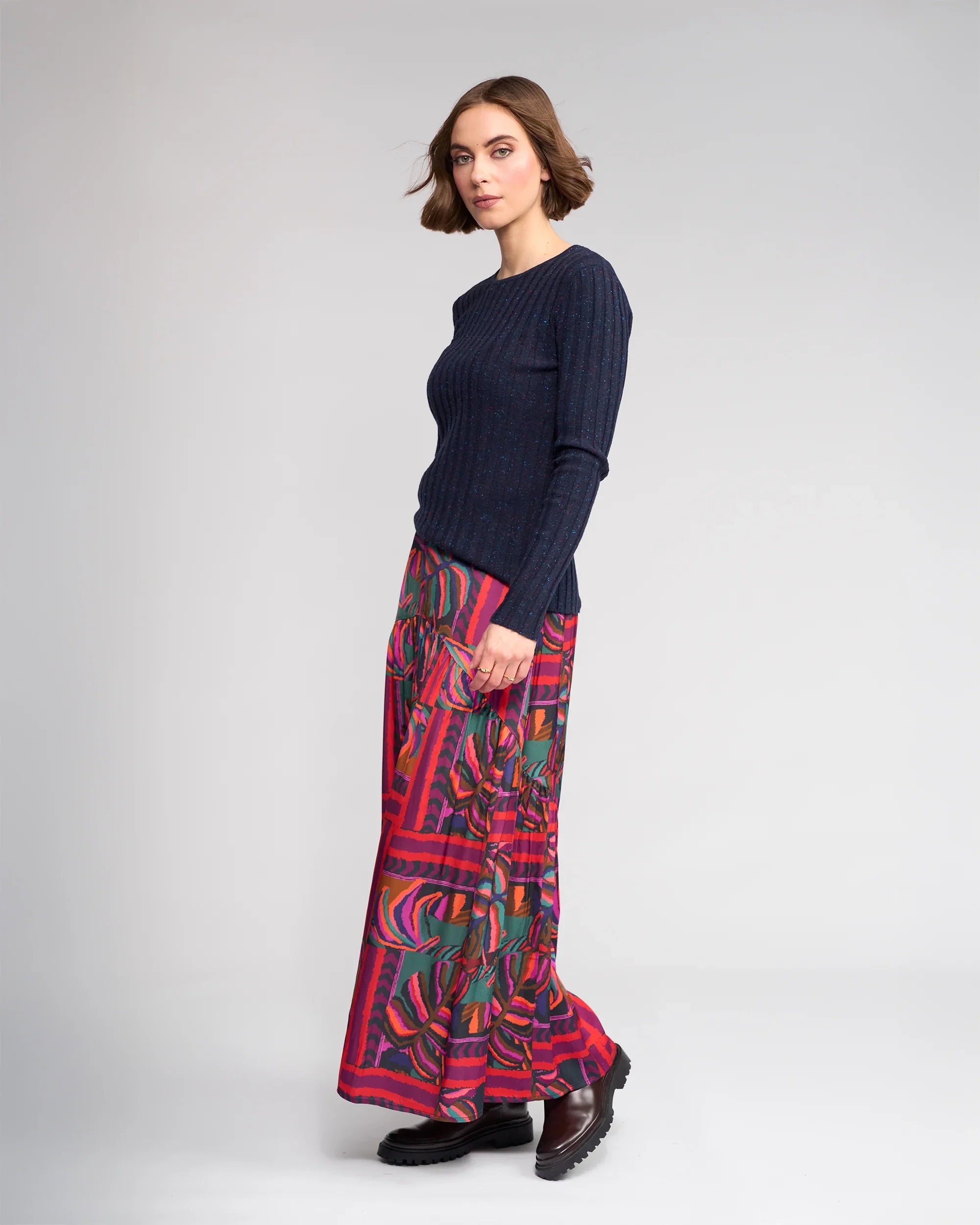 A woman with short brown hair stands sideways against a plain light gray background, wearing a fitted navy ribbed sweater, Knewe Label’s Milan Skirt - Sunset in red and green, and brown chunky shoes.