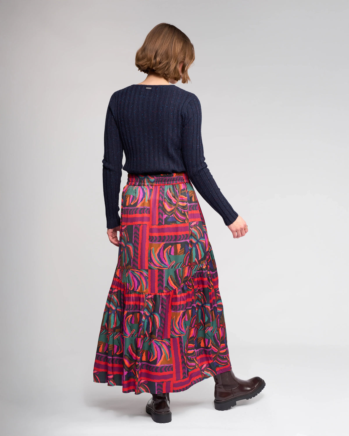 Seen from behind, a person with short brown hair wears a dark ribbed sweater, dark ankle boots, and the Milan Skirt - Sunset by Knewe Label—a flowing full-length skirt with bold geometric and floral patterns—against a plain light background.