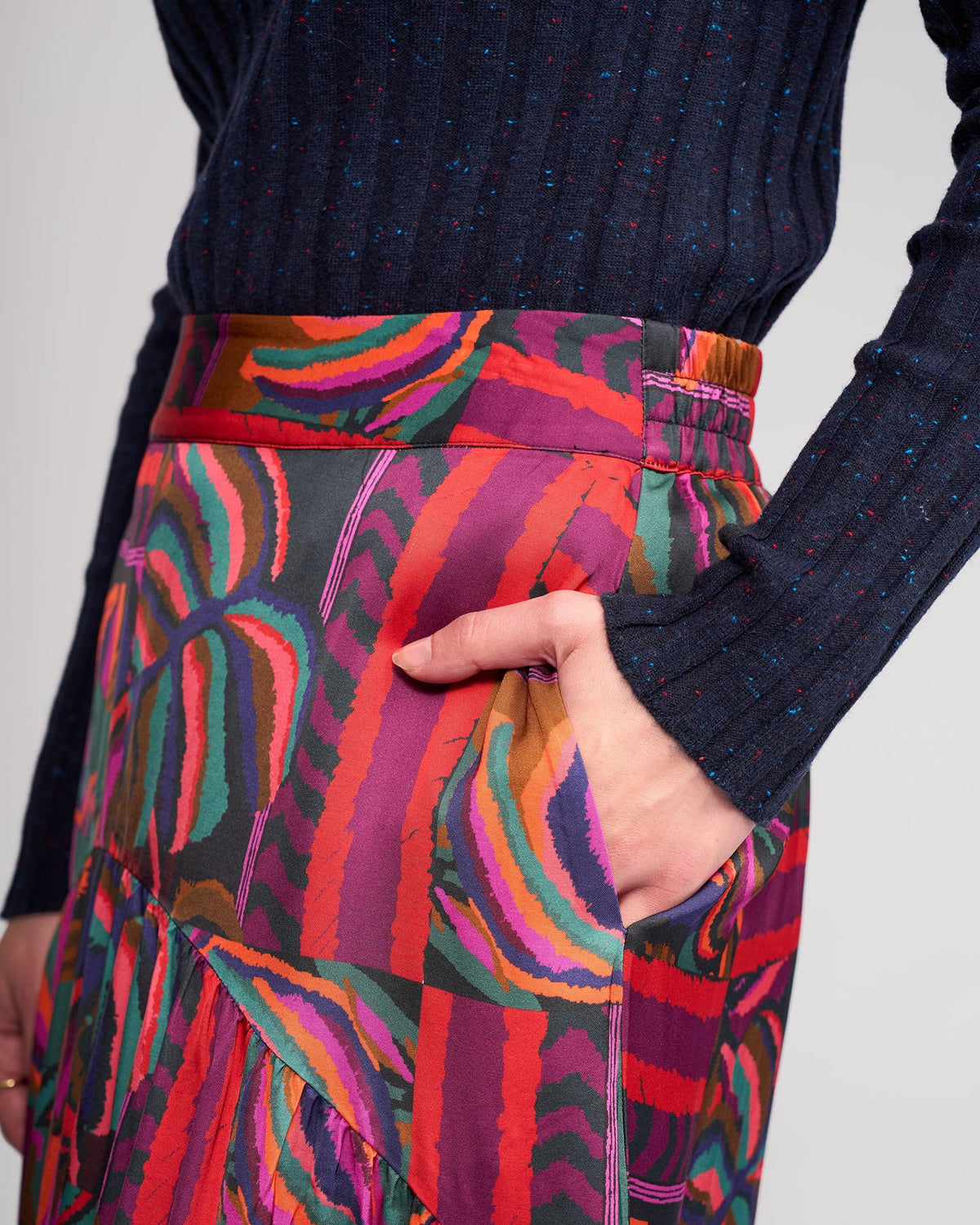 A person wears the Knewe Label Milan Skirt - Sunset, featuring a full-length abstract sunset print, paired with a navy long-sleeve ribbed top, and has one hand in the skirt pocket.