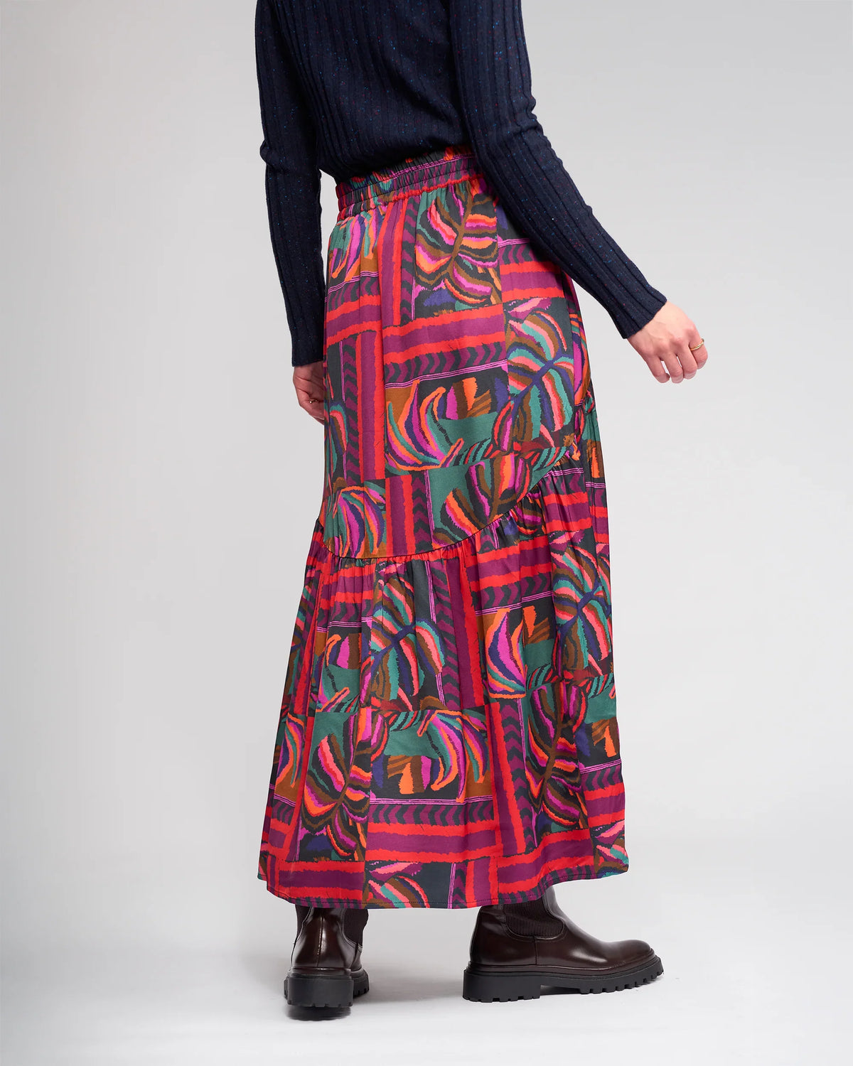 A person wears a dark ribbed sweater, black boots, and the Milan Skirt - Sunset by Knewe Label with red and green leaf designs, standing against a plain light background.