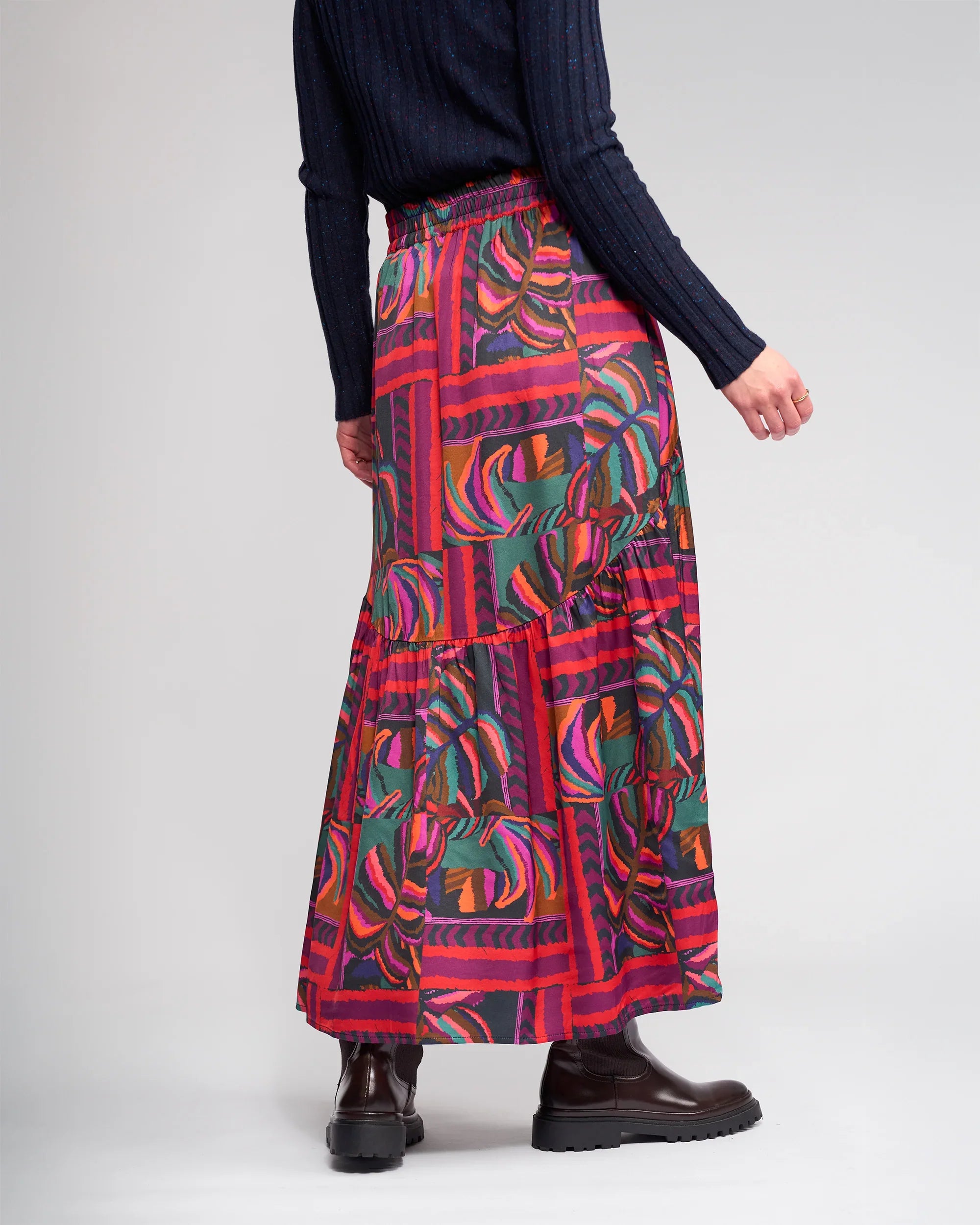 A person wears a dark ribbed sweater, black boots, and the Milan Skirt - Sunset by Knewe Label with red and green leaf designs, standing against a plain light background.