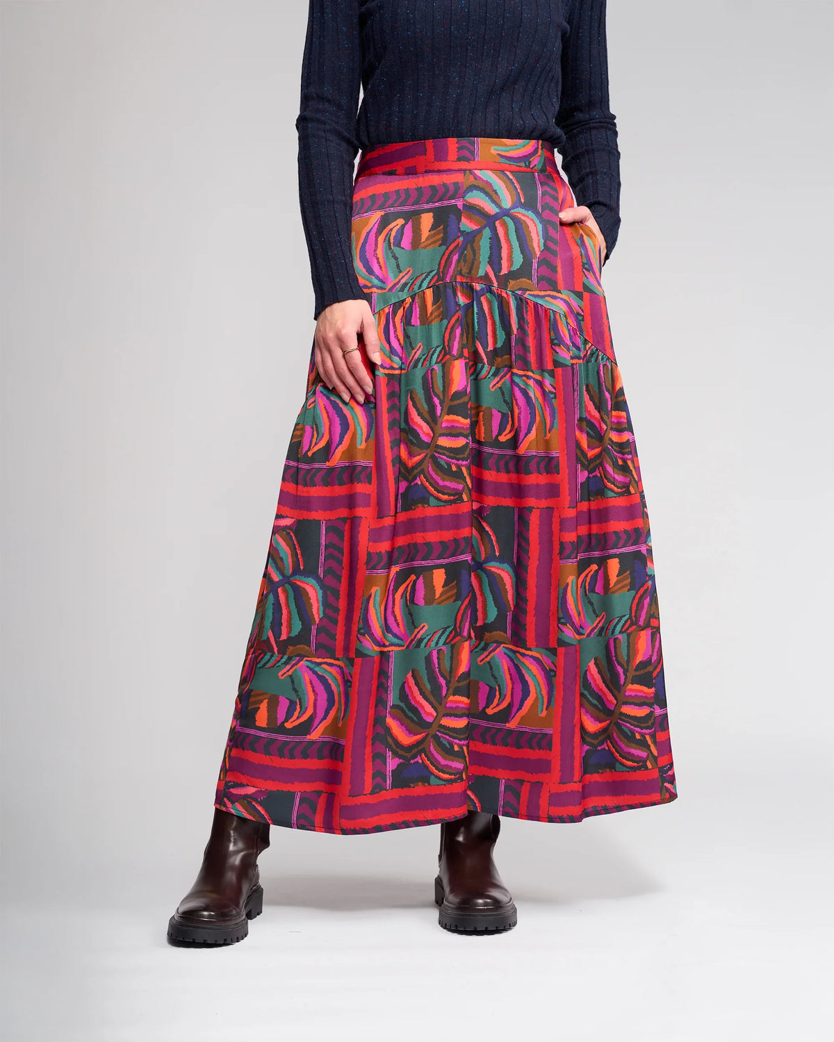 A person wears a dark ribbed long-sleeve top, dark brown ankle boots, and the Knewe Label Milan Skirt - Sunset, a vibrant full-length skirt with abstract red, pink, orange, green, and blue designs. The person's face is not visible.