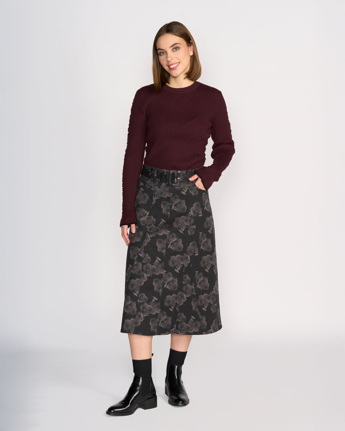 A woman smiles with one hand in her pocket, wearing a dark burgundy sweater and the Anita Skirt - Echo by Knewe Label, a grey floral print A-line denim skirt. She completes her look with black ankle socks and black ankle boots.
