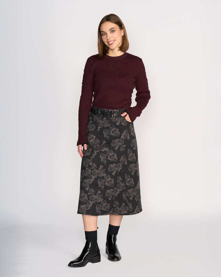 A woman smiles with one hand in her pocket, wearing a dark burgundy sweater and the Anita Skirt - Echo by Knewe Label, a grey floral print A-line denim skirt. She completes her look with black ankle socks and black ankle boots.