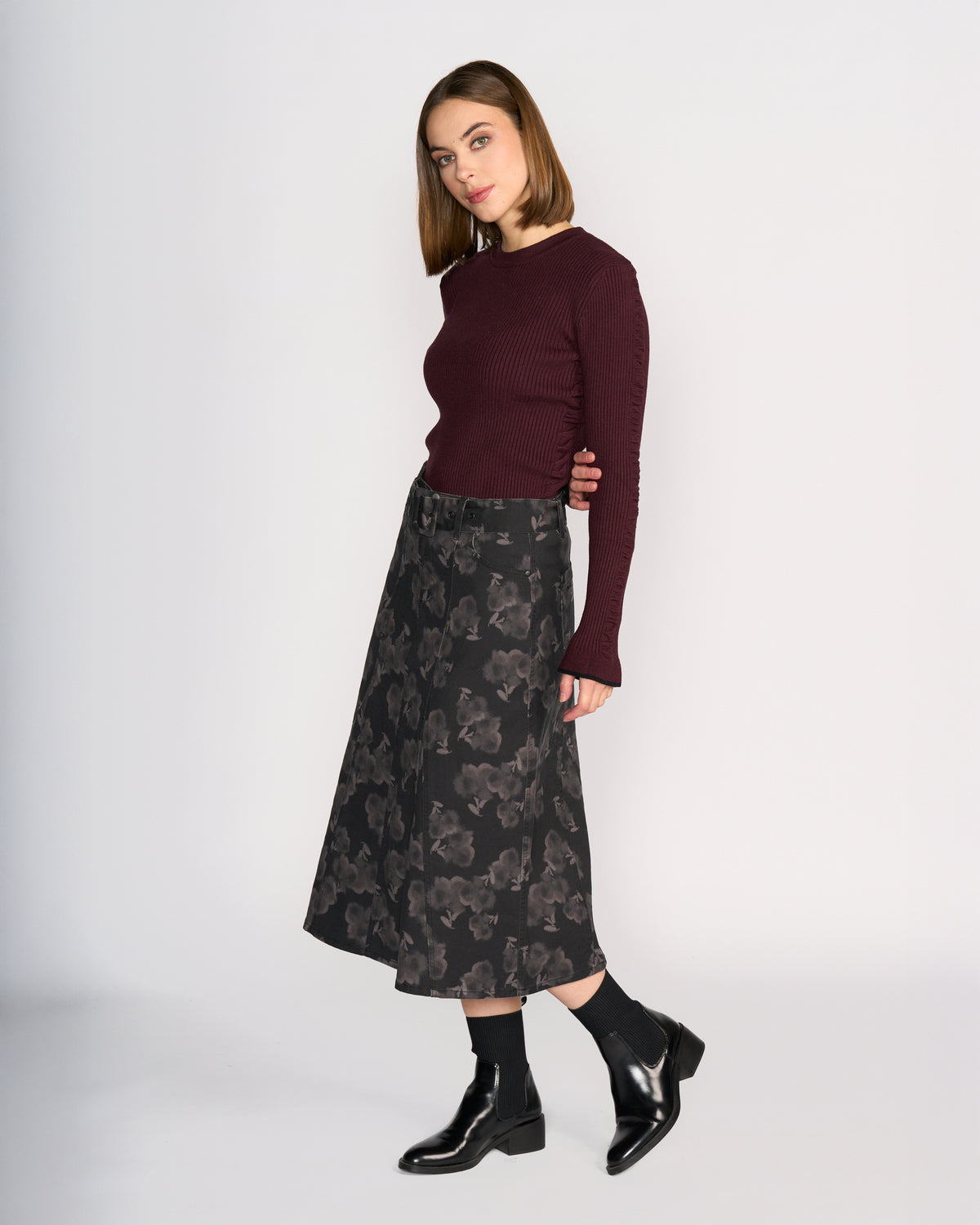 A woman with straight brown hair, wearing a burgundy ribbed sweater, black socks, black ankle boots, and the Anita Skirt - Echo by Knewe Label stands against a plain white background.