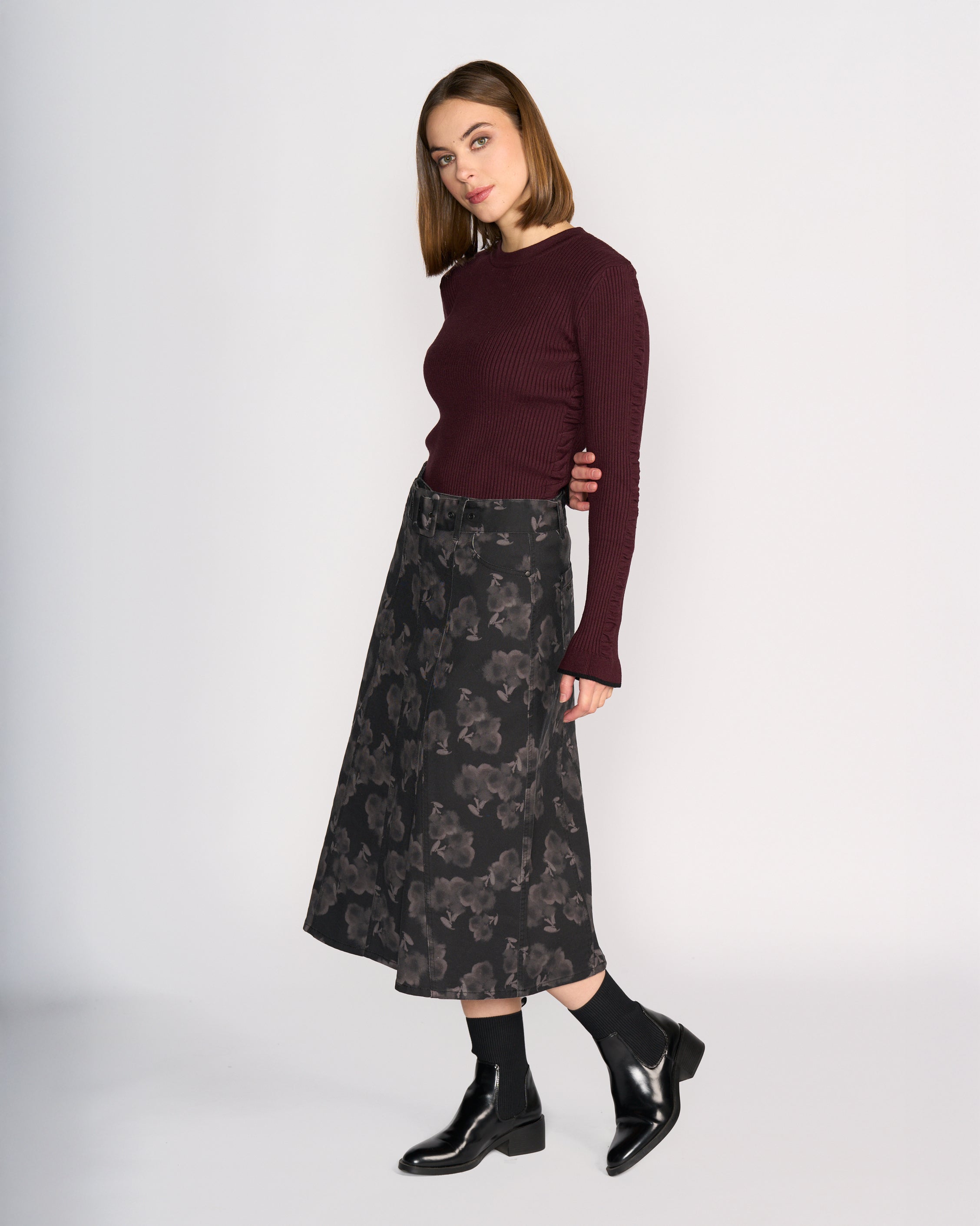 A woman with straight brown hair, wearing a burgundy ribbed sweater, black socks, black ankle boots, and the Anita Skirt - Echo by Knewe Label stands against a plain white background.