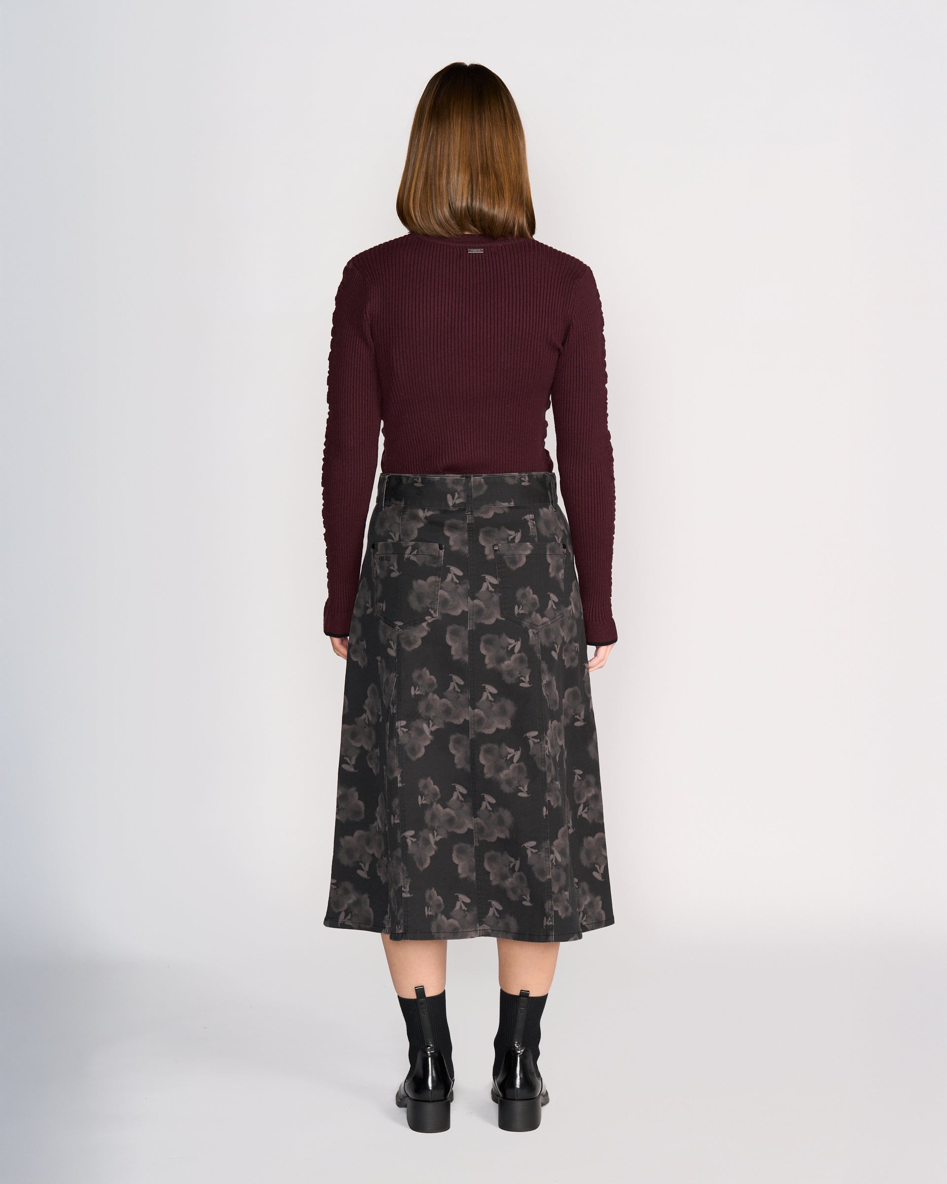 A woman with straight, medium-length brown hair stands facing away against a plain light background, wearing Knewe Label's Anita Skirt - Echo, a maroon long-sleeve top, and black ankle boots.