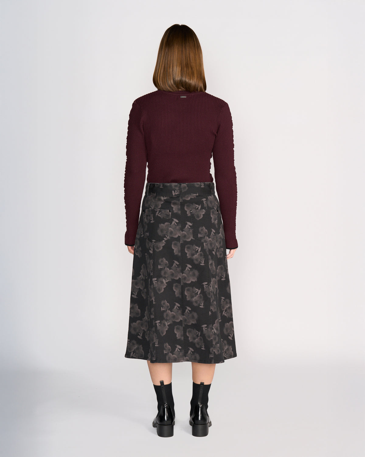 A woman with straight, medium-length brown hair stands facing away against a plain light background, wearing Knewe Label's Anita Skirt - Echo, a maroon long-sleeve top, and black ankle boots.