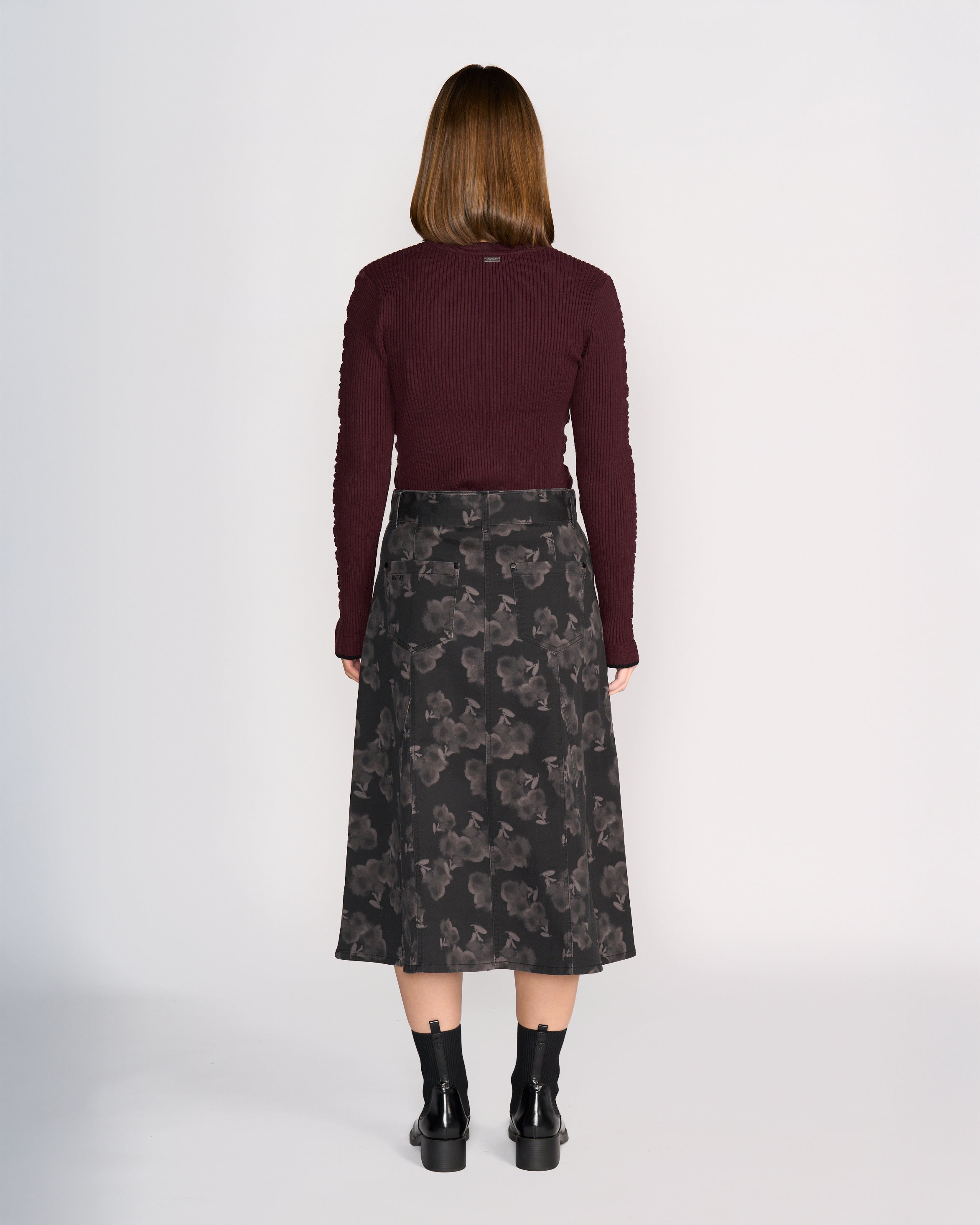 A woman with straight, medium-length brown hair stands facing away against a plain light background, wearing Knewe Label's Anita Skirt - Echo, a maroon long-sleeve top, and black ankle boots.