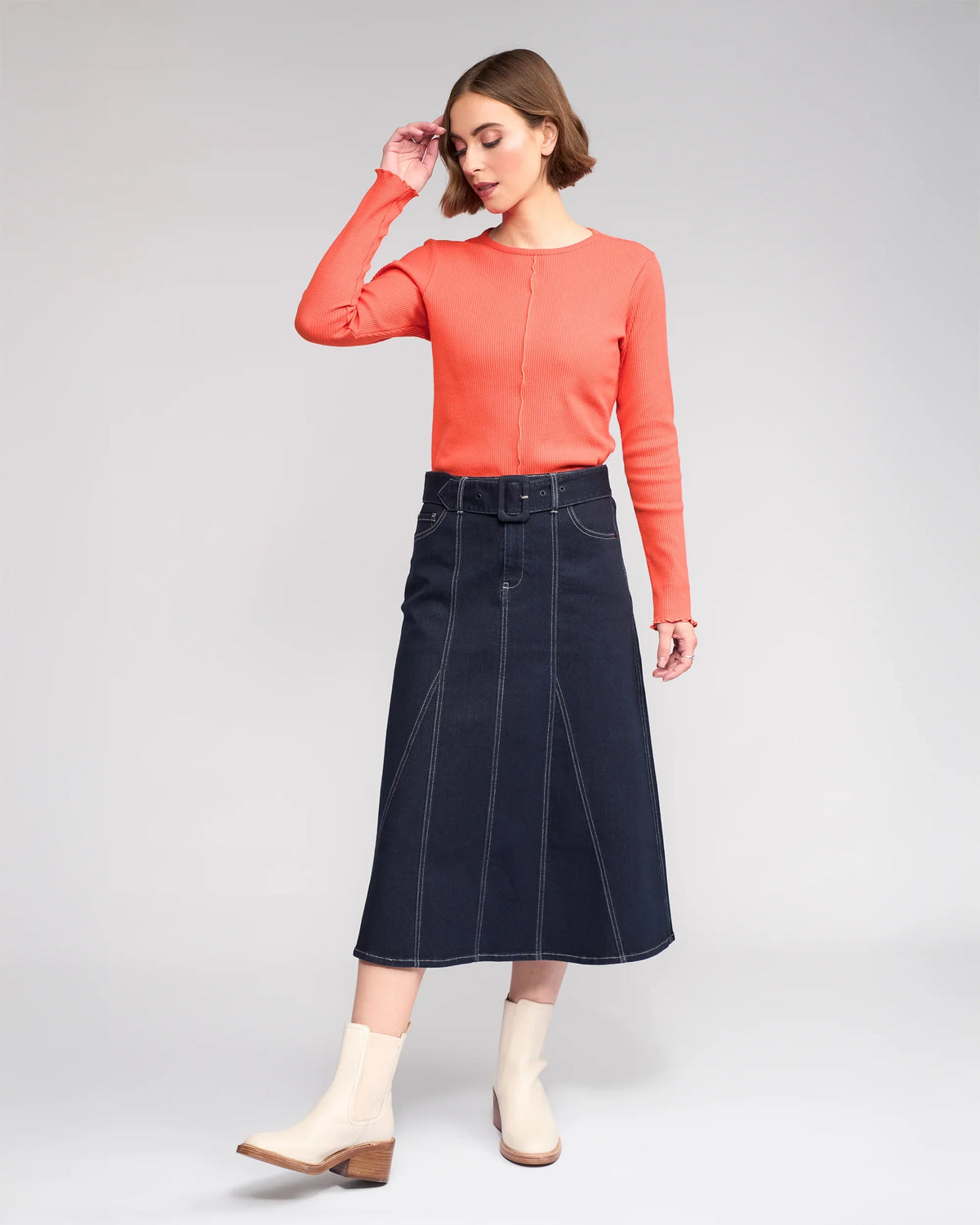 A woman poses in a coral long-sleeve top and the Anita Skirt in Ink Denim by Knewe Label, featuring visible stitching and a denim belt, styled with white ankle boots with low wooden heels against a plain light background.
