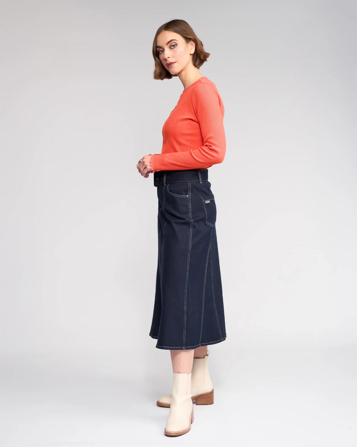 A woman with short brown hair and a neutral expression stands sideways against a plain backdrop, wearing an orange long-sleeve top, light ankle boots, and the Knewe Label Anita Skirt in Ink Denim with a denim belt.