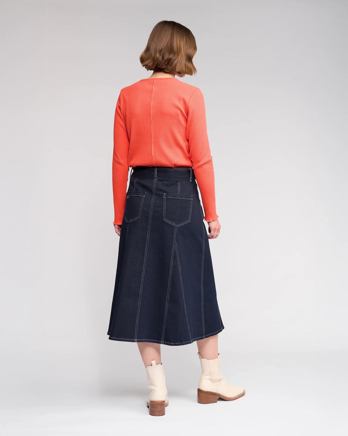 A person with short brown hair faces away, wearing a bright orange long-sleeve top, Knewe Label's Anita Skirt in Ink Denim with visible stitching and back pockets, and cream ankle boots against a plain background.
