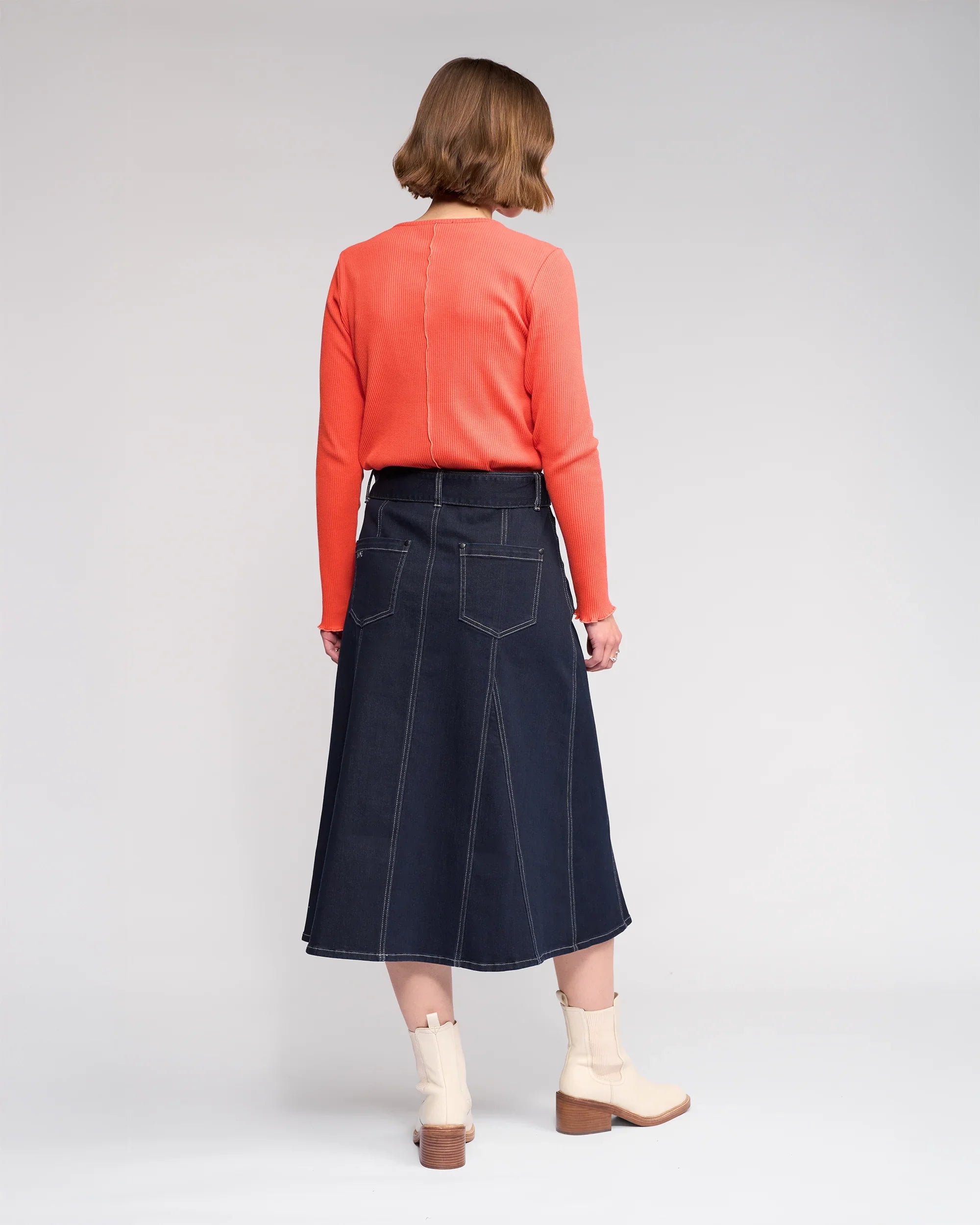 A person with short brown hair faces away, wearing a bright orange long-sleeve top, Knewe Label's Anita Skirt in Ink Denim with visible stitching and back pockets, and cream ankle boots against a plain background.