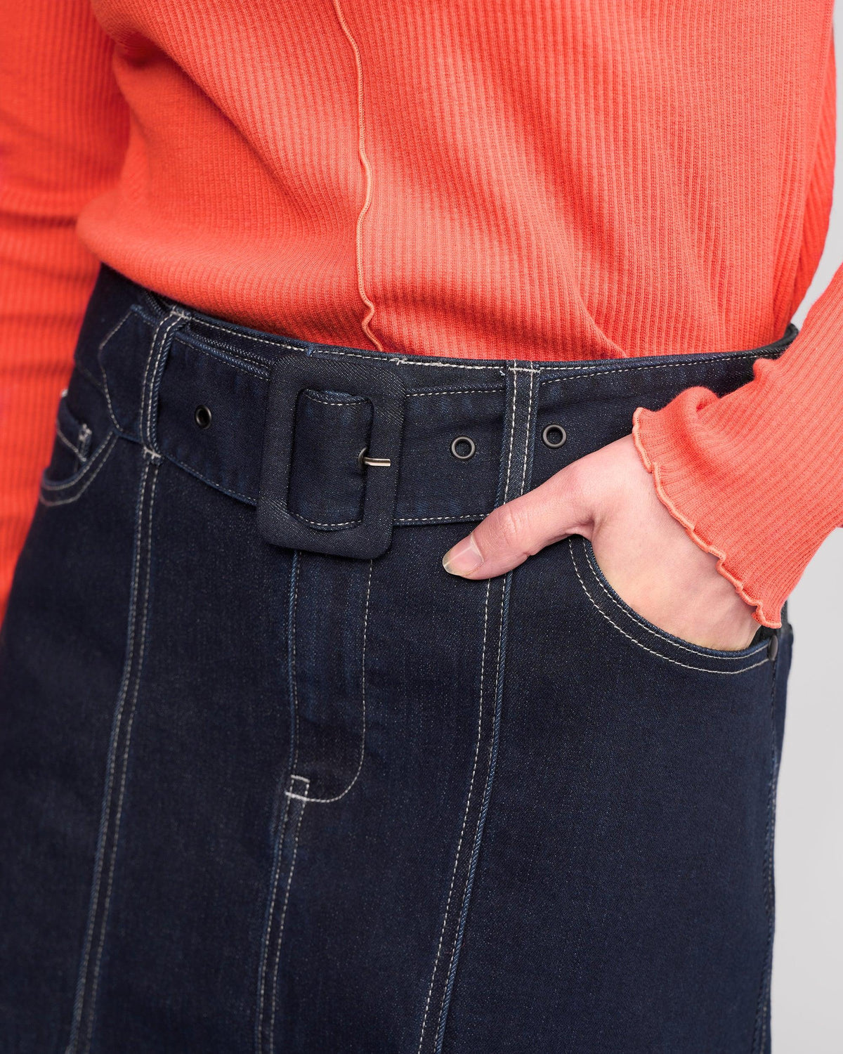 A person wears a ribbed, long-sleeve coral top with ruffled cuffs and the Anita Skirt - Ink Denim by Knewe Label, featuring a denim belt and white stitching, standing with one hand in their pocket.