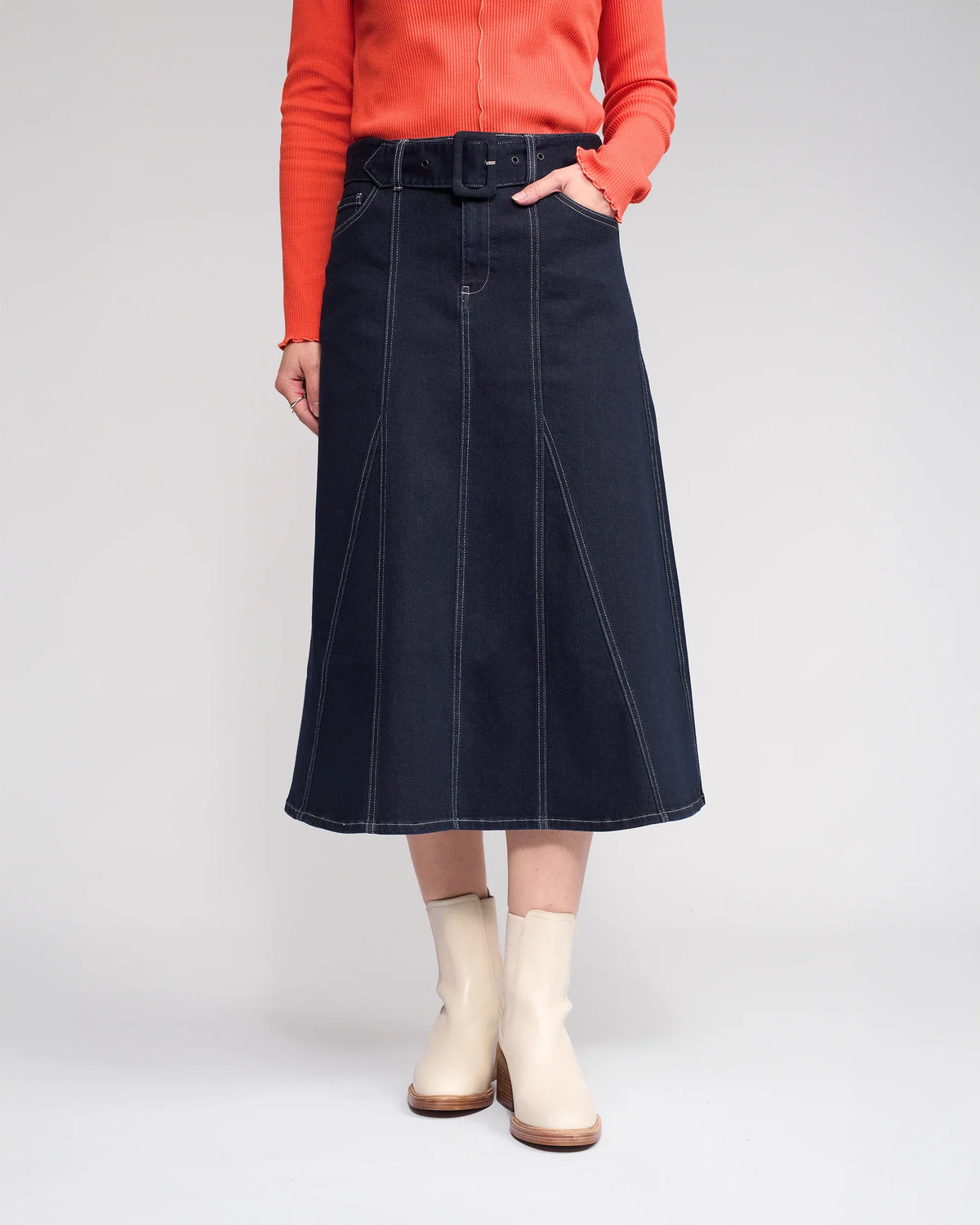 A person wearing an orange long-sleeve top, the Knewe Label Anita Skirt in Ink Denim with a denim belt, and light beige ankle boots stands against a plain light background.