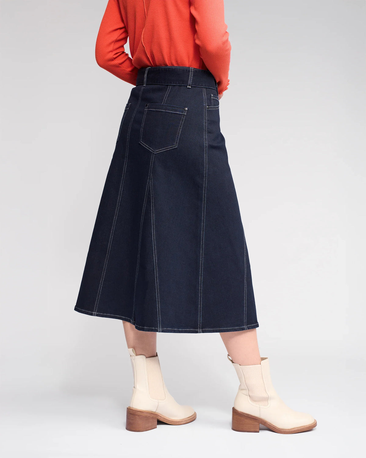A person wears an orange long-sleeve top, Knewe Label’s Anita Skirt in Ink Denim with a matching belt and white stitching, plus light beige ankle boots with a wooden block heel, standing against a plain light background.