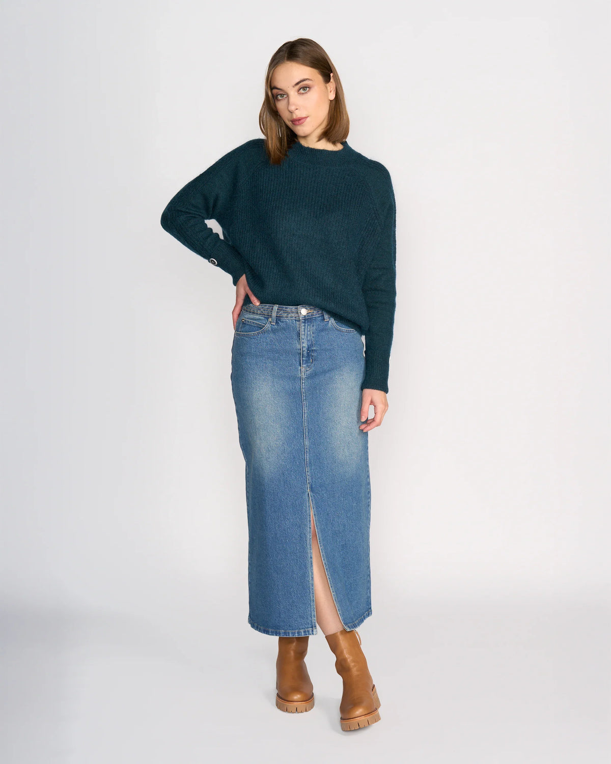 A woman with straight, shoulder-length hair stands in a relaxed pose against a plain background, wearing the Knewe Label Palmer Skirt - Vintage Denim in a mid-blue wash with a front slit, paired with a dark green sweater and tan heeled boots.