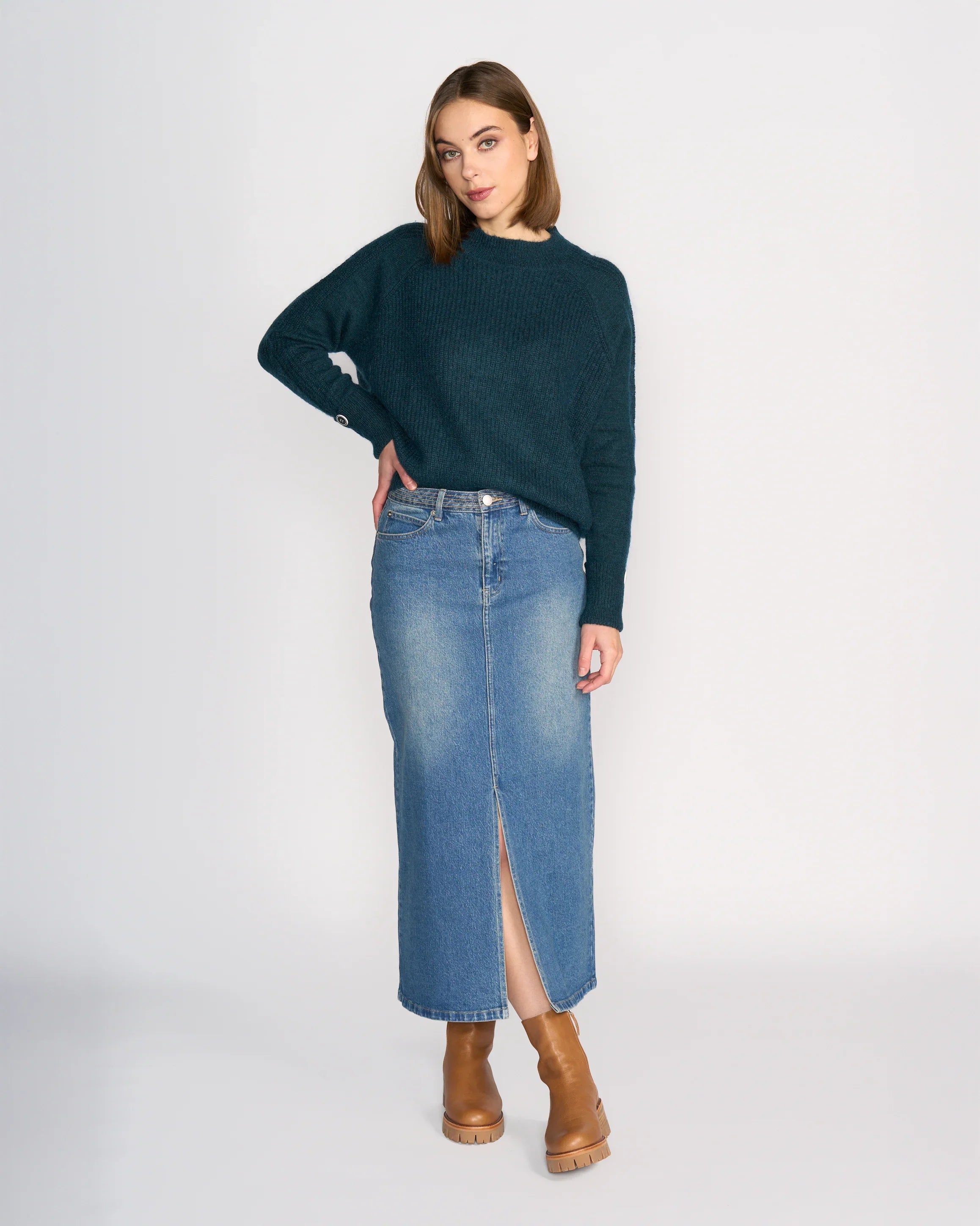 A woman with straight, shoulder-length hair stands in a relaxed pose against a plain background, wearing the Knewe Label Palmer Skirt - Vintage Denim in a mid-blue wash with a front slit, paired with a dark green sweater and tan heeled boots.