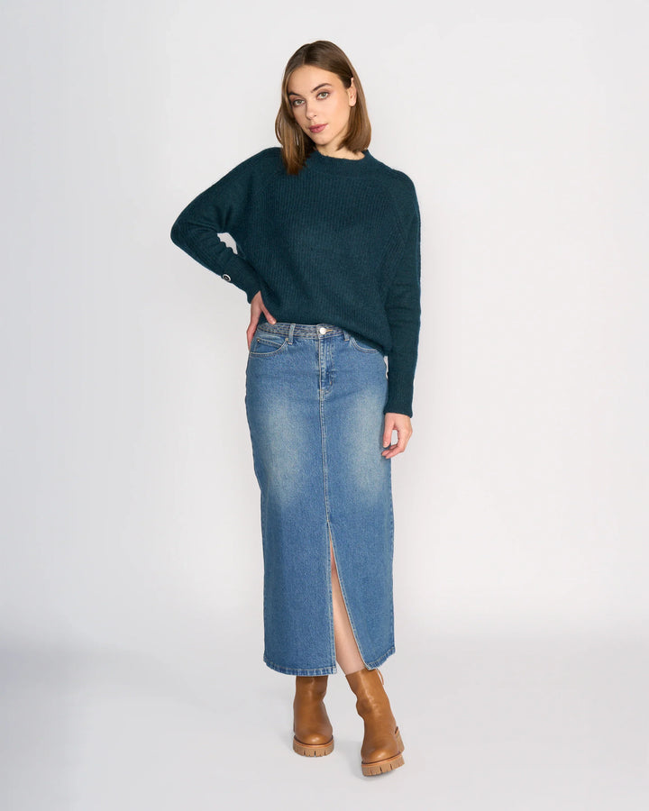 A woman with straight, shoulder-length hair stands in a relaxed pose against a plain background, wearing the Knewe Label Palmer Skirt - Vintage Denim in a mid-blue wash with a front slit, paired with a dark green sweater and tan heeled boots.