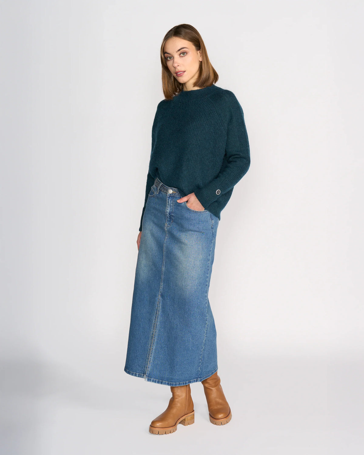 A woman with shoulder-length brown hair stands against a plain background, wearing Knewe Label's Palmer Skirt in Vintage Denim, a dark green sweater, and tan ankle boots while looking slightly to the side.