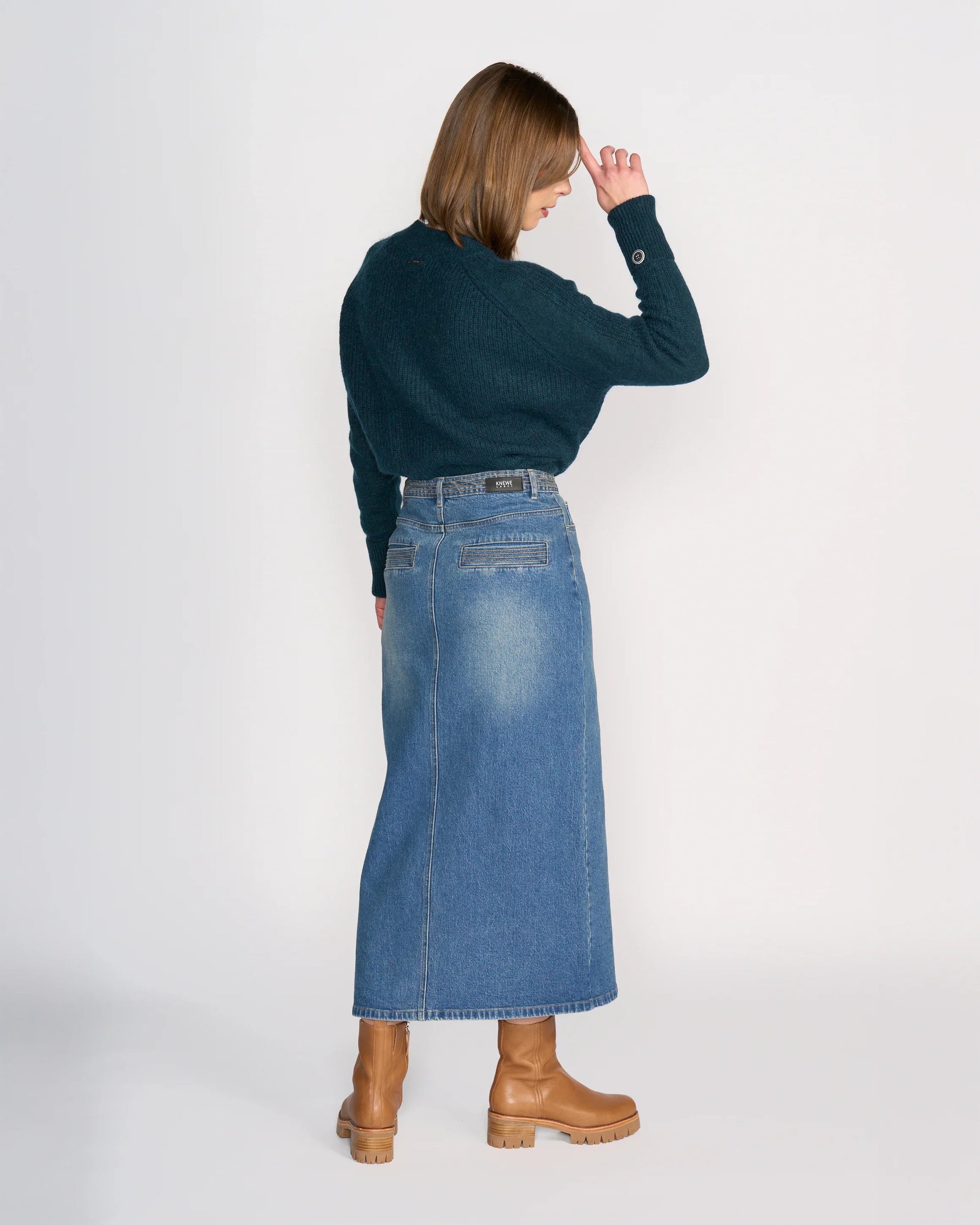 A woman, with brown hair down, stands against a white background wearing the Knewe Label Palmer Skirt in vintage denim, a dark green sweater, and tan heeled boots. She faces away with one hand to her forehead.