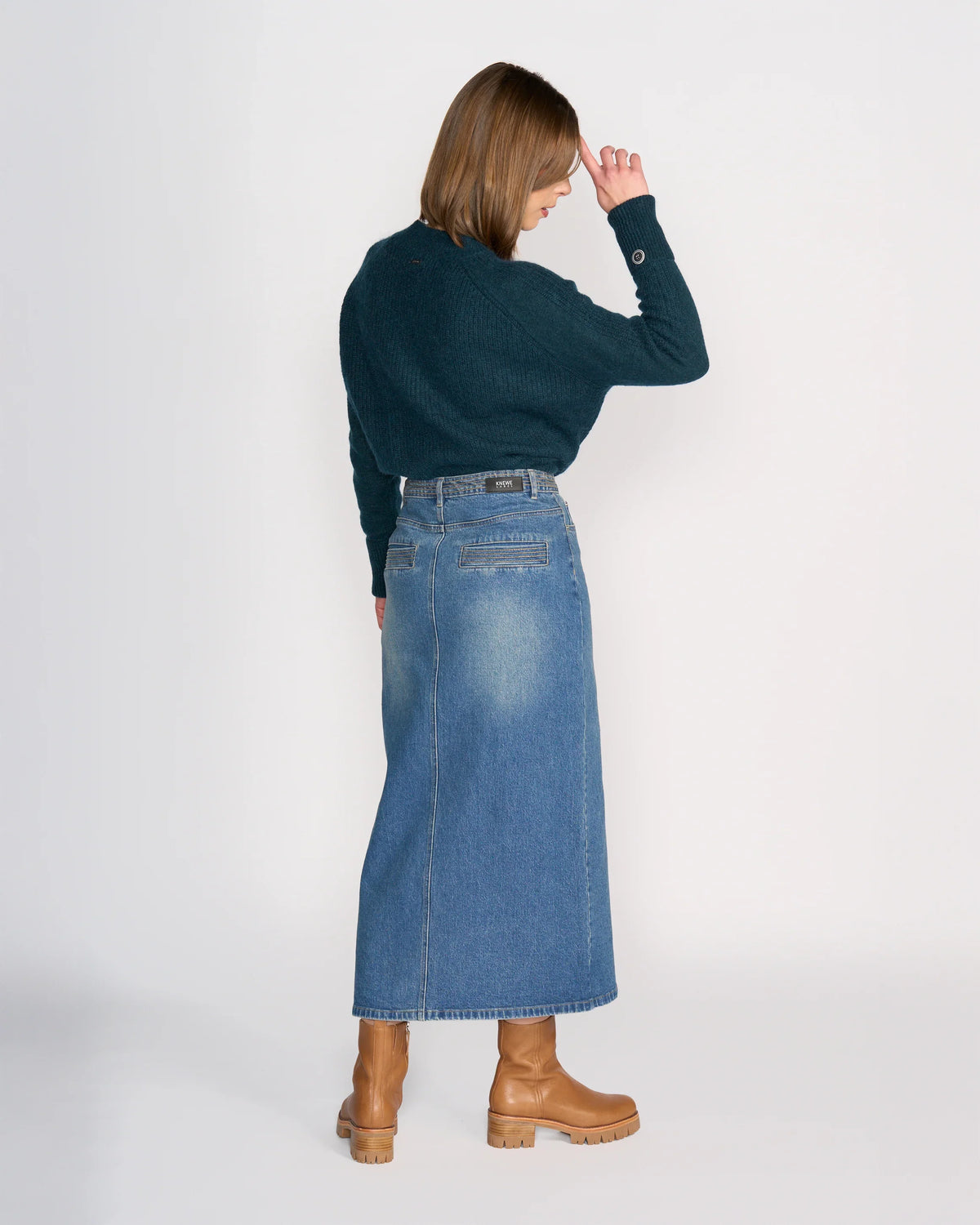A woman, with brown hair down, stands against a white background wearing the Knewe Label Palmer Skirt in vintage denim, a dark green sweater, and tan heeled boots. She faces away with one hand to her forehead.