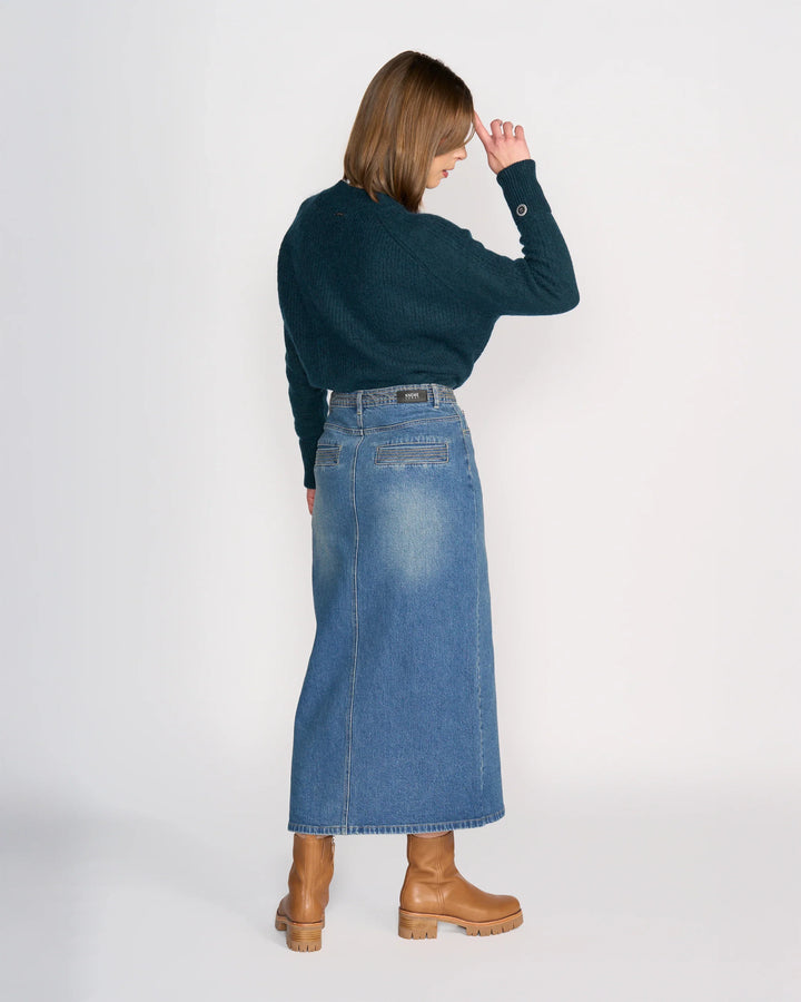 A woman, with brown hair down, stands against a white background wearing the Knewe Label Palmer Skirt in vintage denim, a dark green sweater, and tan heeled boots. She faces away with one hand to her forehead.