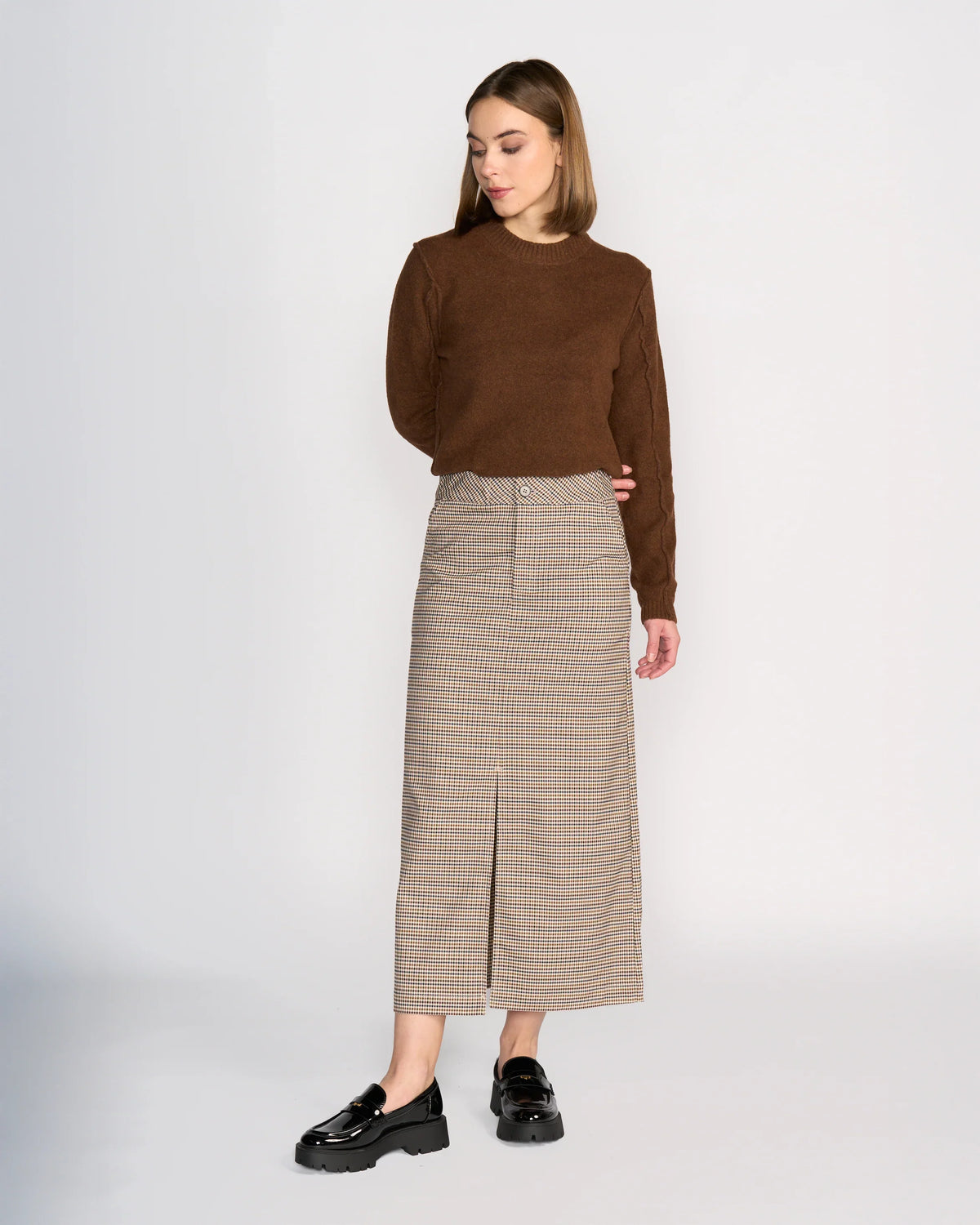 A woman stands against a plain background wearing Knewe Label’s Rhodes Skirt - Paige, a structured maxi with a flattering silhouette and front slit, paired with a brown sweater and black chunky loafers. She looks down with her hands behind her back.