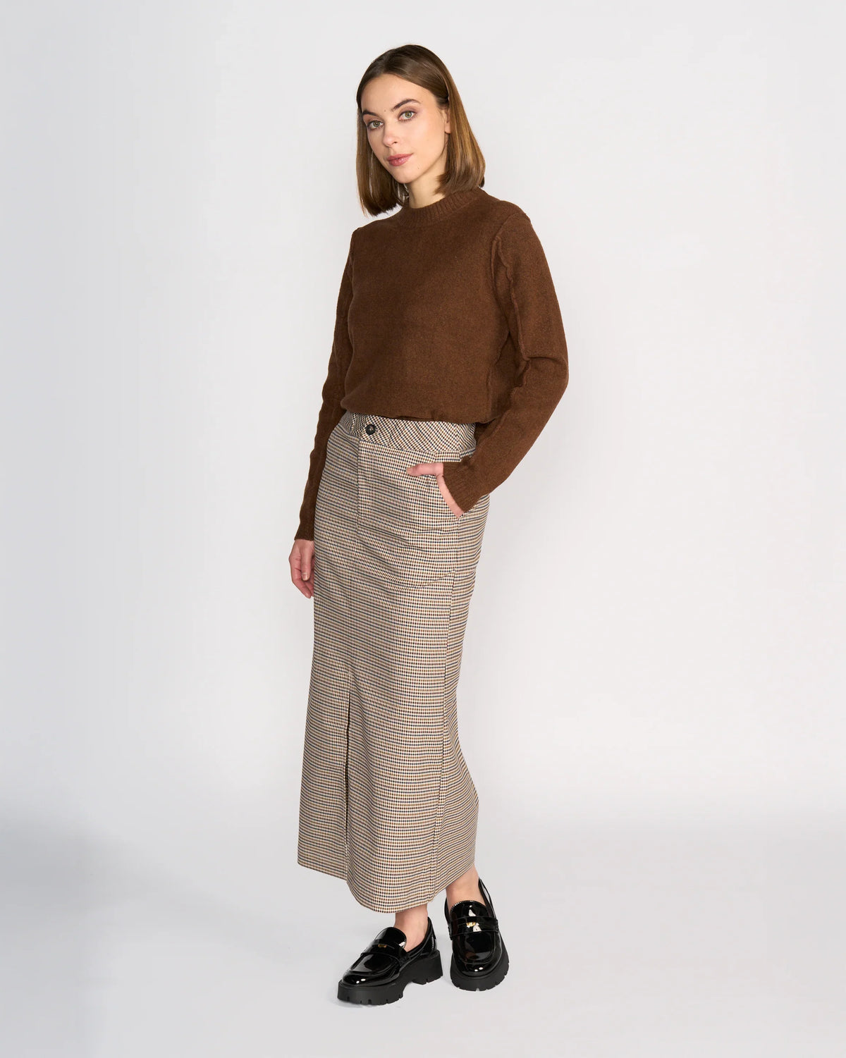 A woman stands against a plain background wearing a brown long-sleeve top, black loafers, and the Knewe Label Rhodes Skirt in the Paige plaid—a structured maxi skirt that flatters her silhouette as she poses with one hand in her pocket.