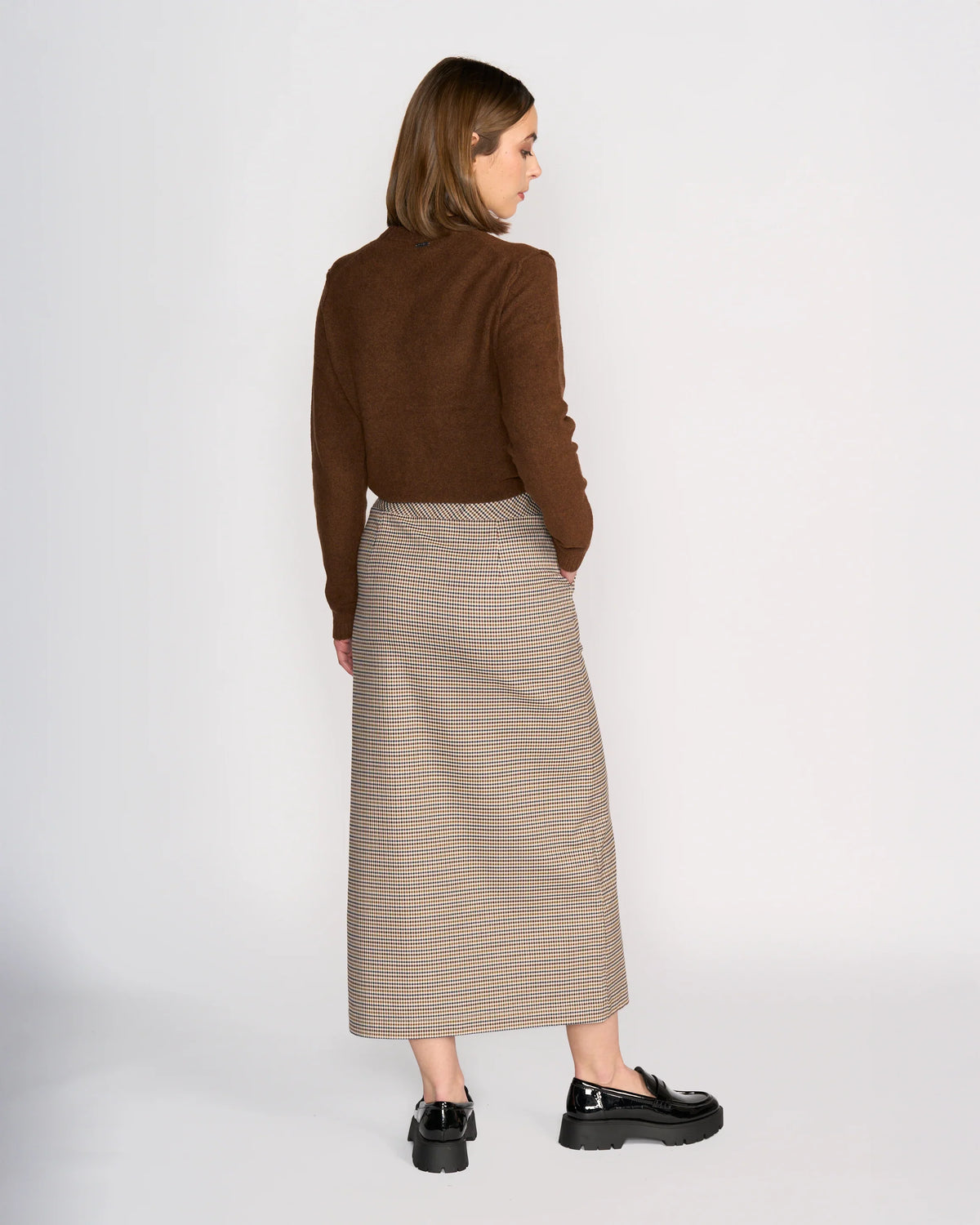 A woman with straight, shoulder-length hair faces away from the camera against a light background, wearing the Knewe Label Rhodes Skirt - Paige with a brown long-sleeve top and black loafers, creating a flattering silhouette.