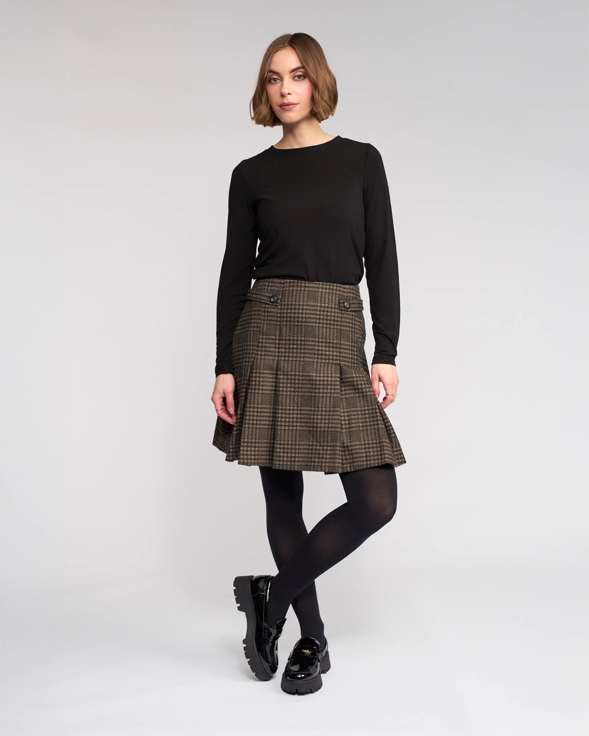 A woman with a short bob stands against a plain background wearing the Knewe Label Soho Skirt - Check, a brown check pleated winter skirt, paired with a black long-sleeve top, black tights, and shoes. Her hands are relaxed at her sides.