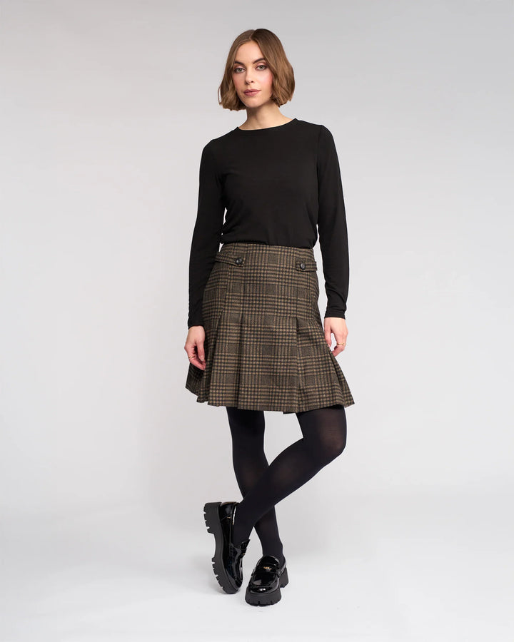 A woman with a short bob stands against a plain background wearing the Knewe Label Soho Skirt - Check, a brown check pleated winter skirt, paired with a black long-sleeve top, black tights, and shoes. Her hands are relaxed at her sides.