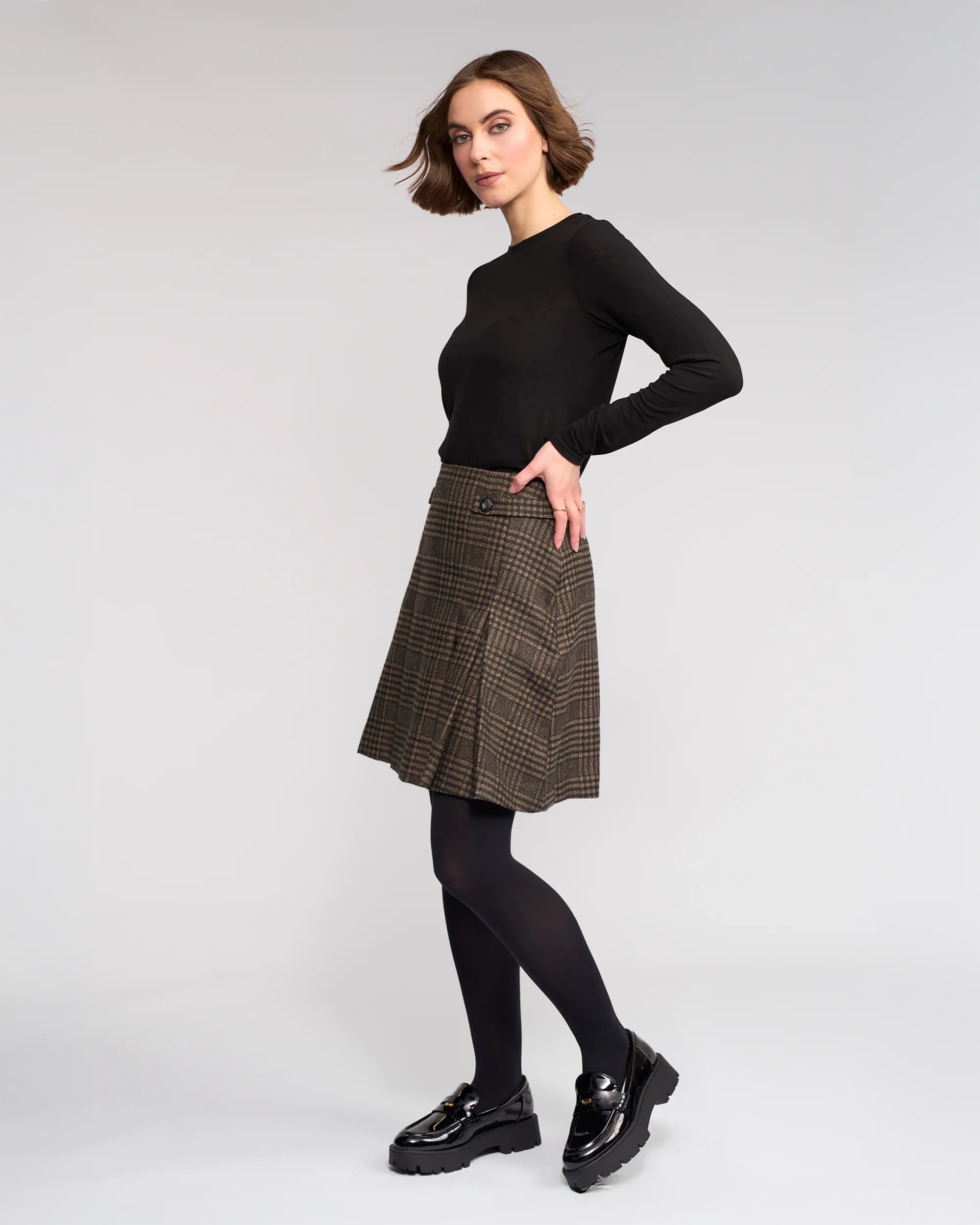A woman stands sideways against a plain light background, looking at the camera with a slight smile, wearing the Soho Skirt - Check by Knewe Label—a pleated brown winter-weight check skirt—paired with a black long-sleeve top, tights, and shoes.