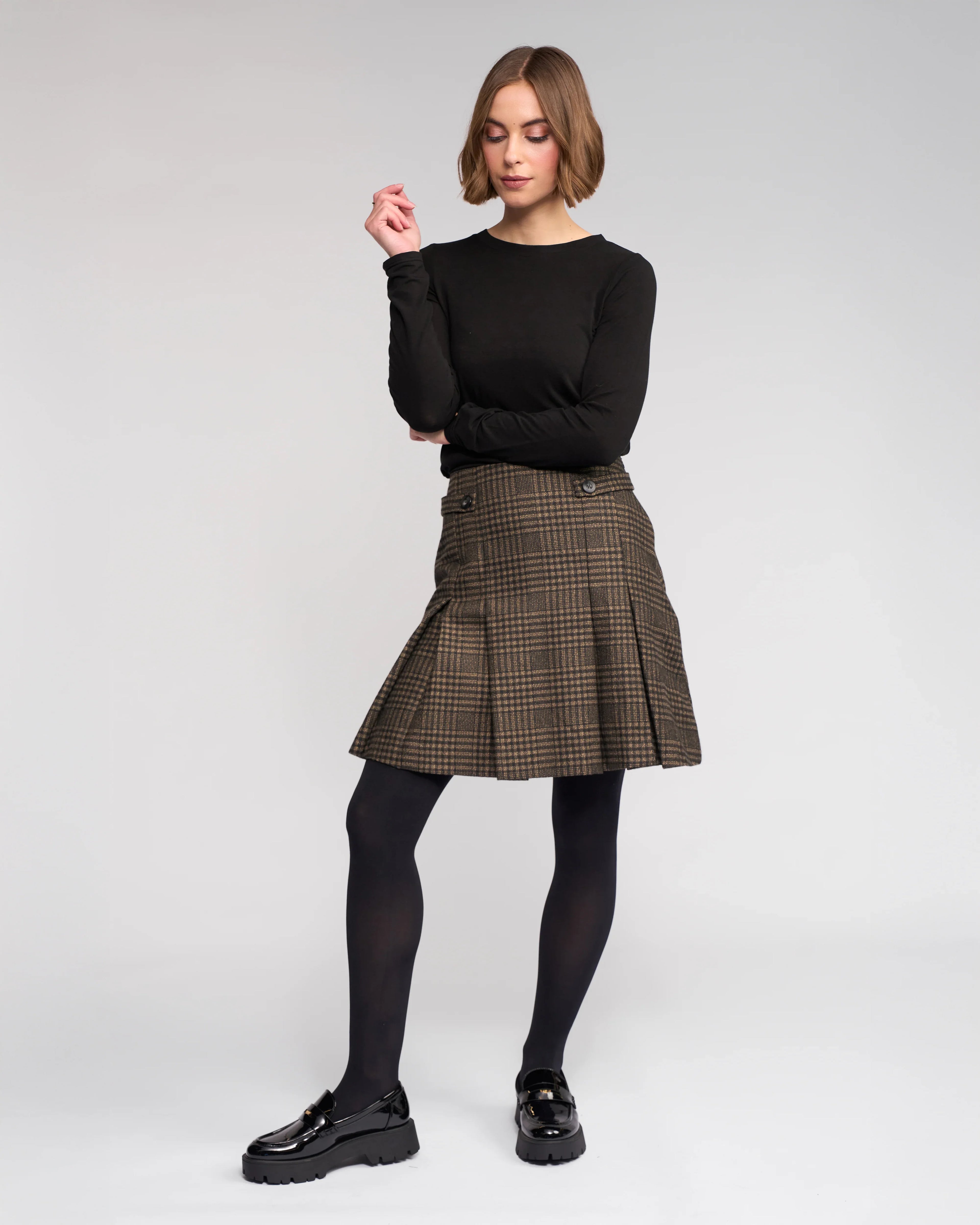 A person stands against a plain light background, wearing the Knewe Label Soho Skirt - Check, a black long-sleeve top, black tights, and loafers. They have short light brown hair, look to the side, and have one arm raised.