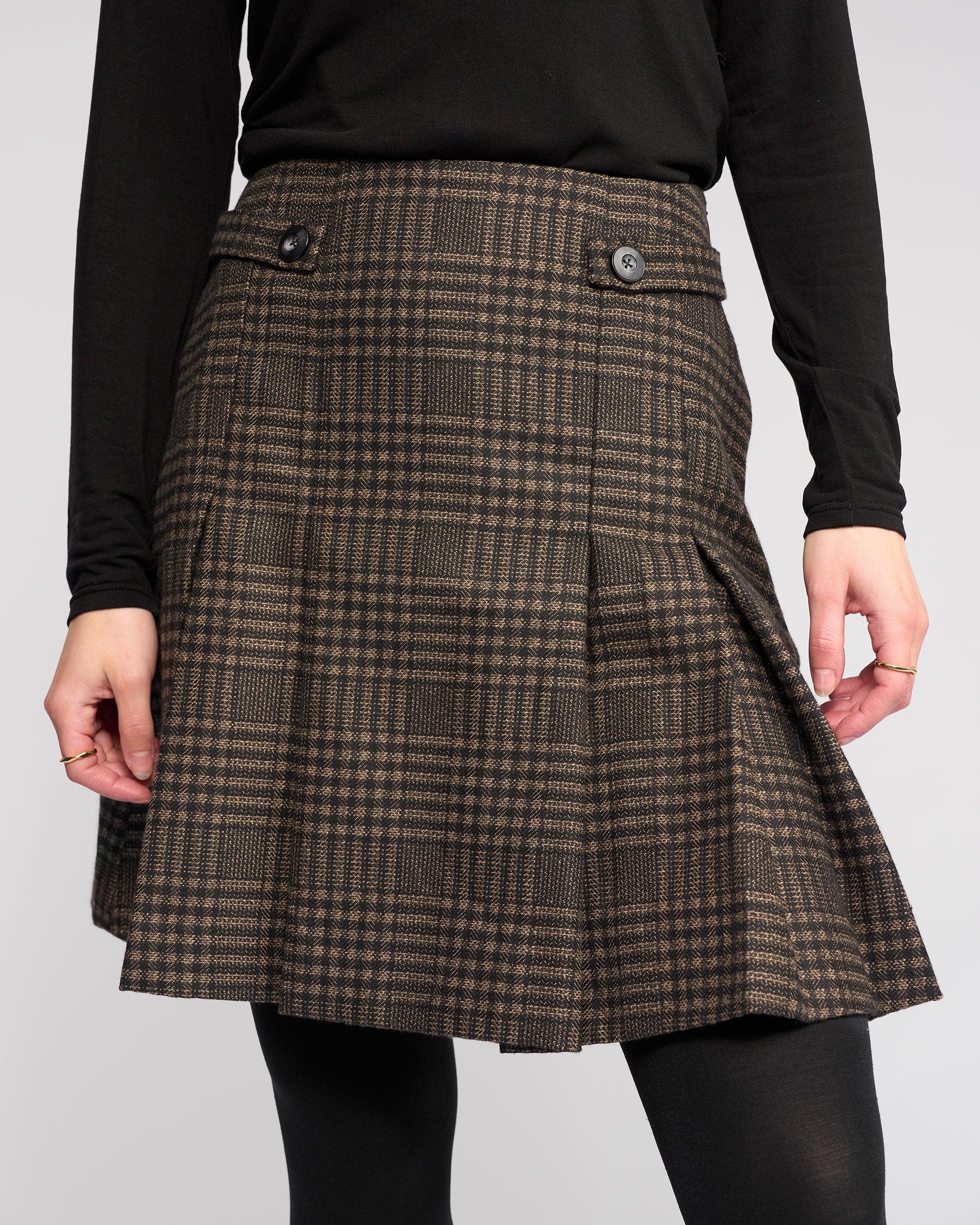 A model wears the Knewe Label Soho Skirt - Check, a brown and black checked pleated winter skirt with waist buttons, styled with a long-sleeve black top and black tights against a plain light background.